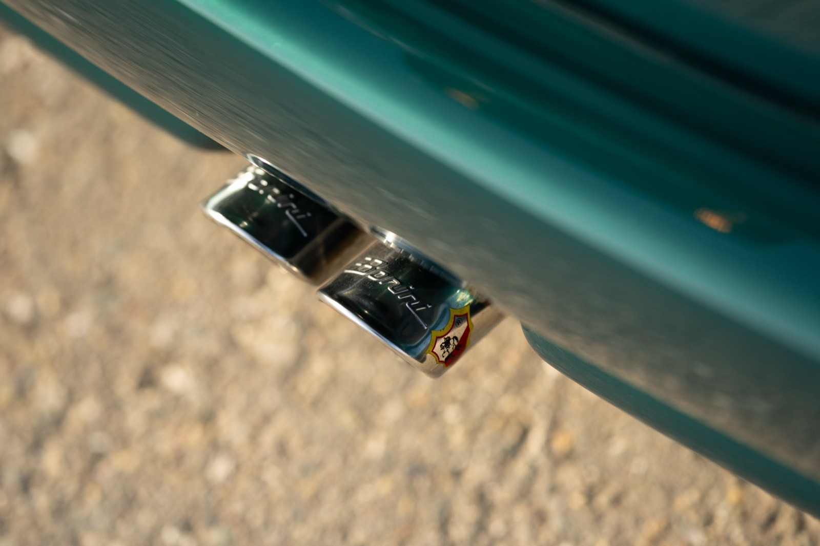 Close-up of a vehicle's exhaust pipe situated on sandy terrain, showcasing its metallic finish and attachment.