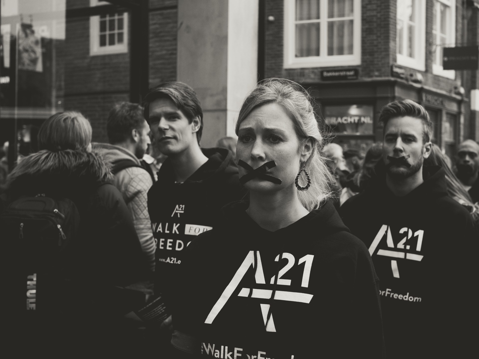 Black-and-white photo of a group of demonstrators wearing black hoodies printed “A21” and “Walk For Freedom,” some with black tape across their mouths, marching down a city street past brick storefronts and shop signs in a solemn anti‑trafficking awareness protest.