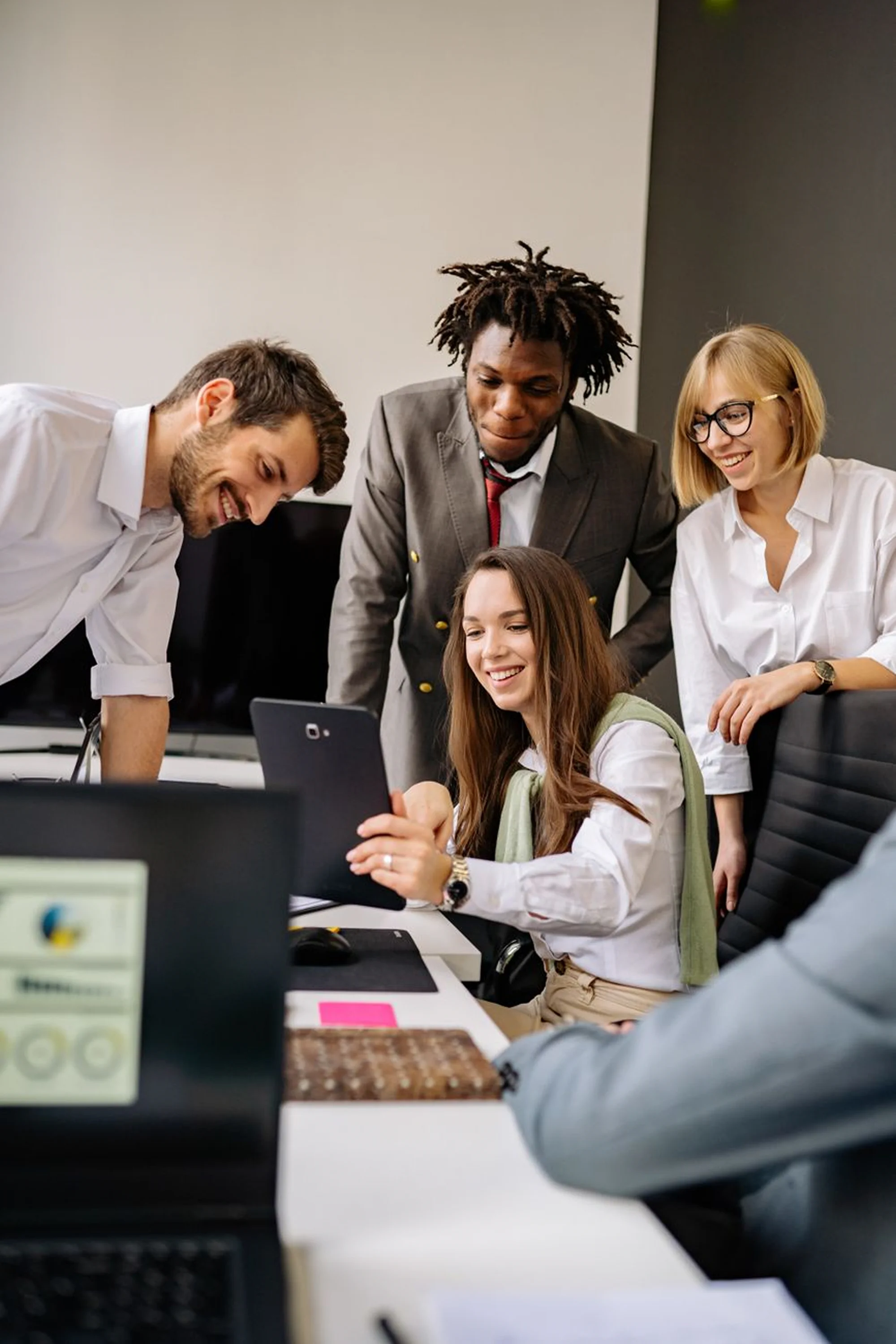 A diverse group of four colleagues gathers around a desk in a modern office, smiling as they look at a tablet held by a seated woman.