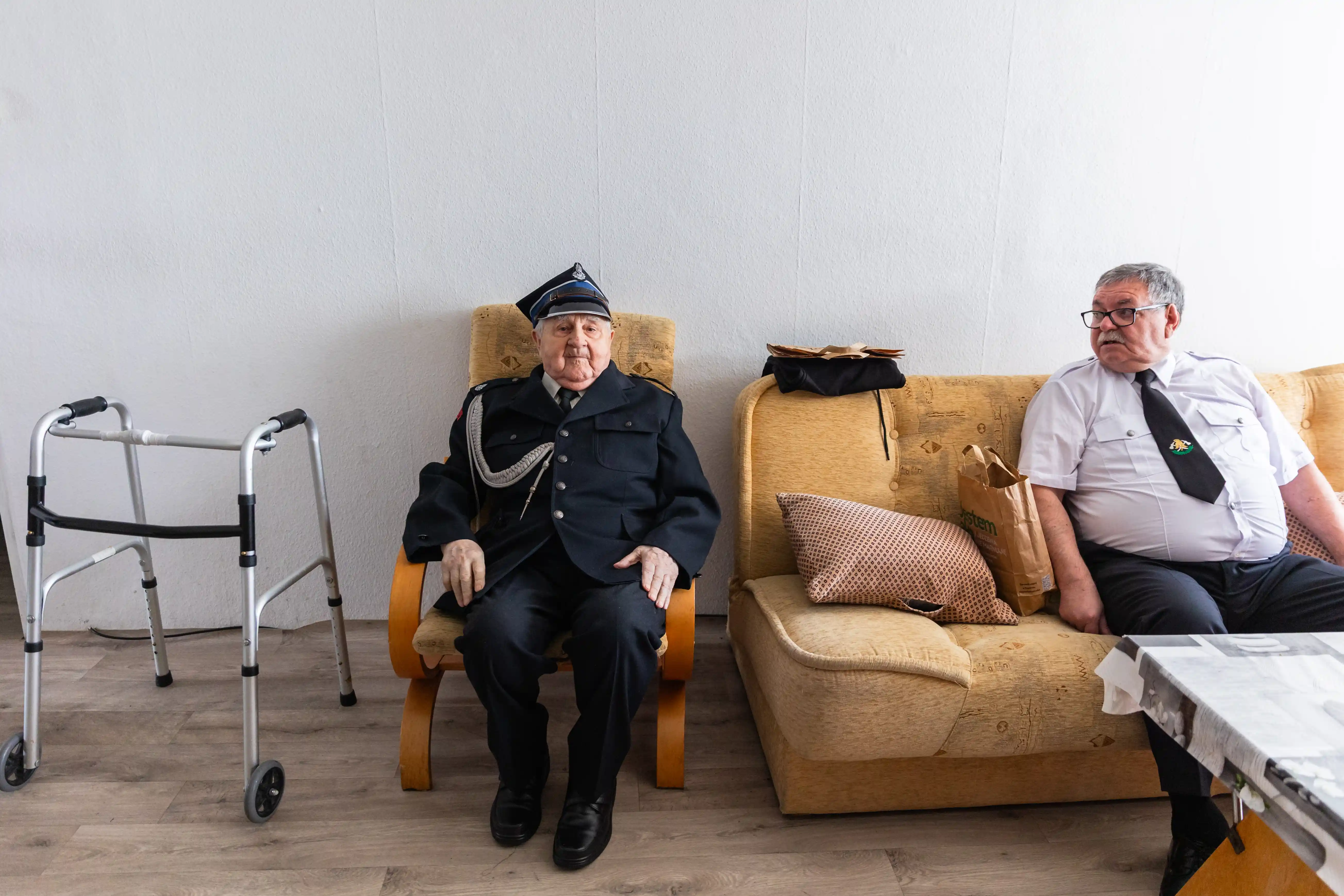 Starszy mężczyzna w mundurze funkcjonariusza siedzi na fotelu, obok niego siedzi na kanapie mężczyzna w białej koszuli i krawacie. Po lewej stronie znajduje się chodzik.