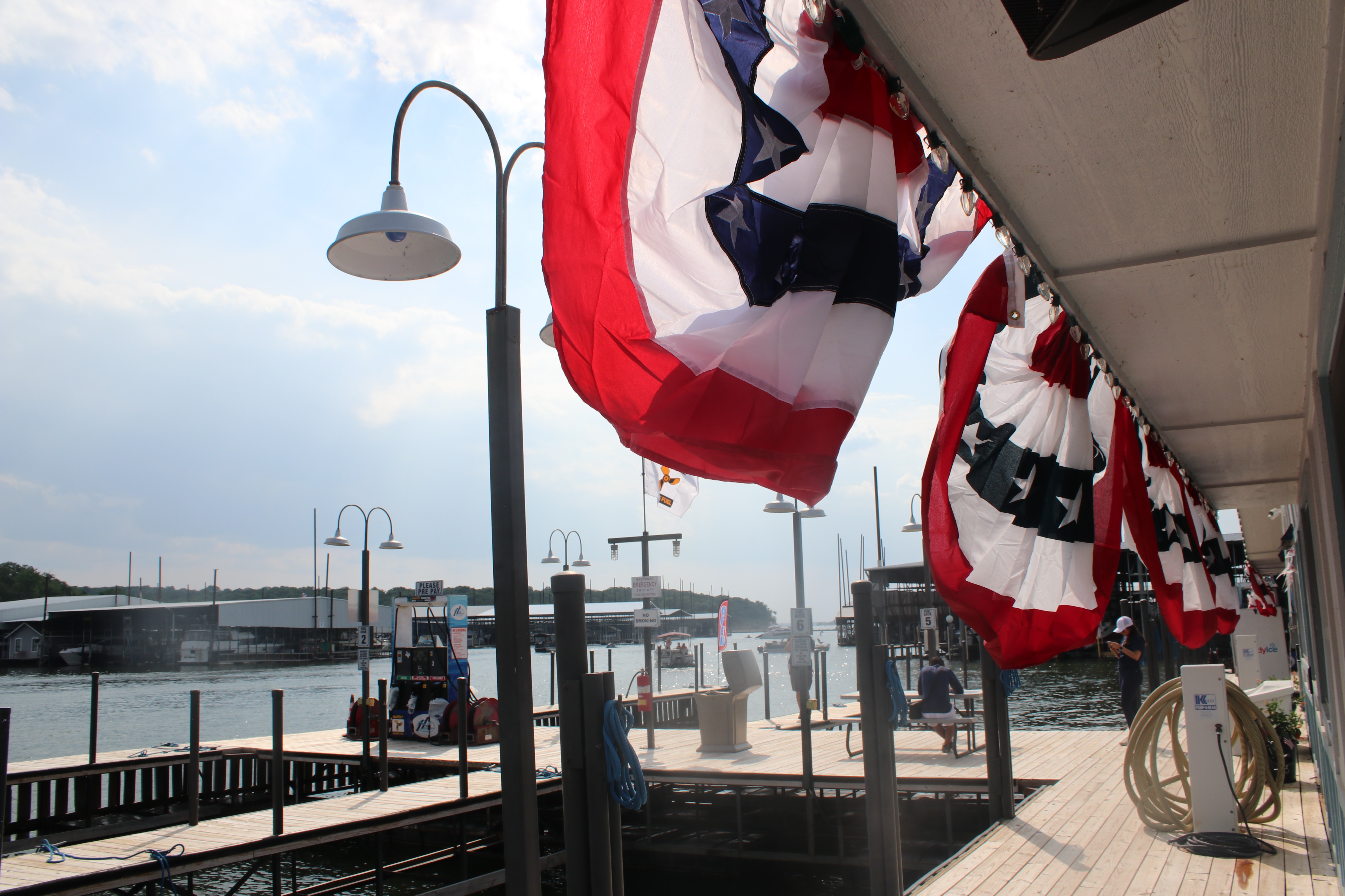 Row of patriotic red, white, and blue bunting flags line the waterfront walkway at a marina, with wooden docks and boats visible against a backdrop of calm water and a partly cloudy sky, enhancing the festive atmosphere.