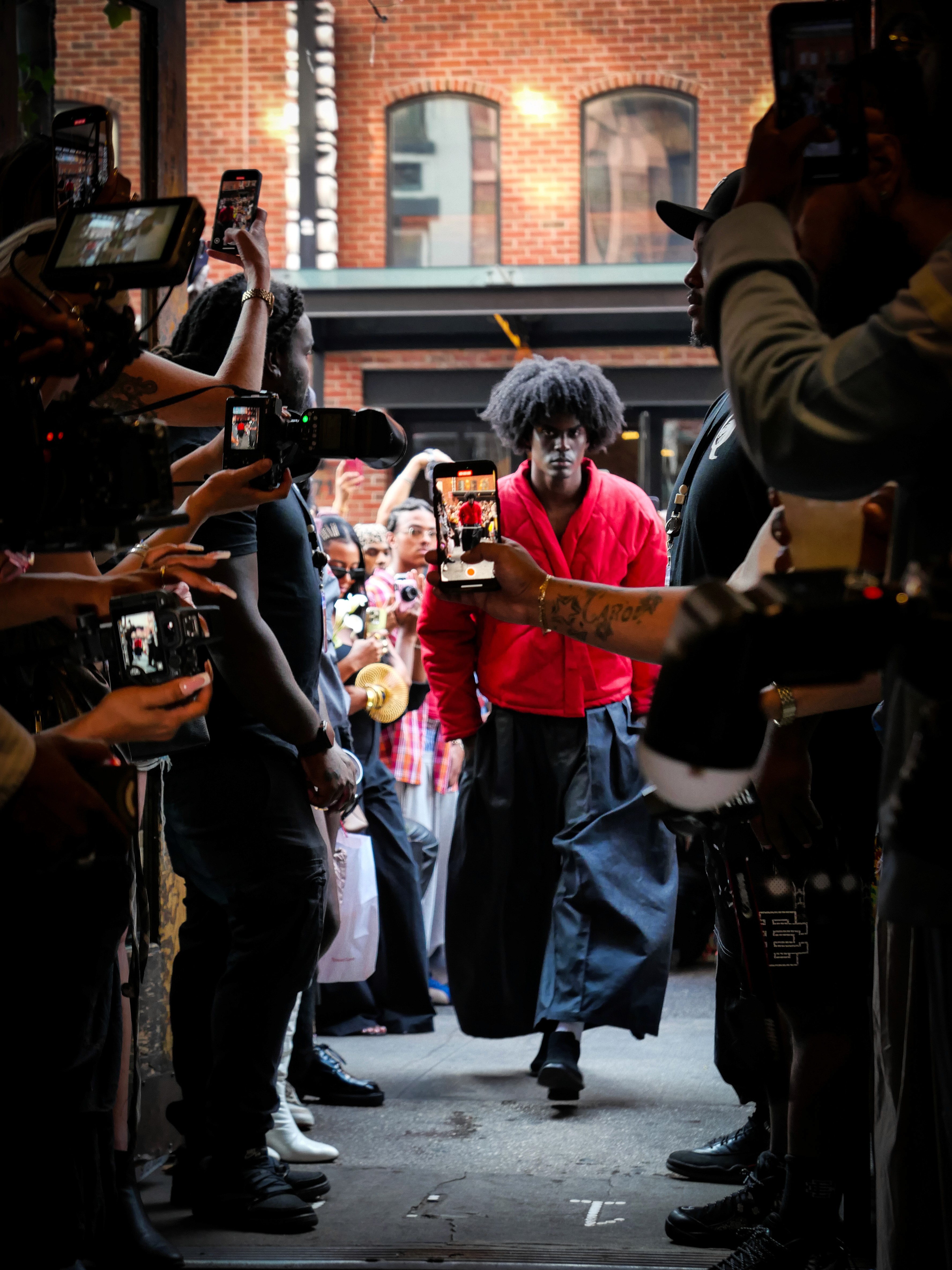 Runway-style entrance shot with photographers framing a model in a red jacket walking through the crowd.
