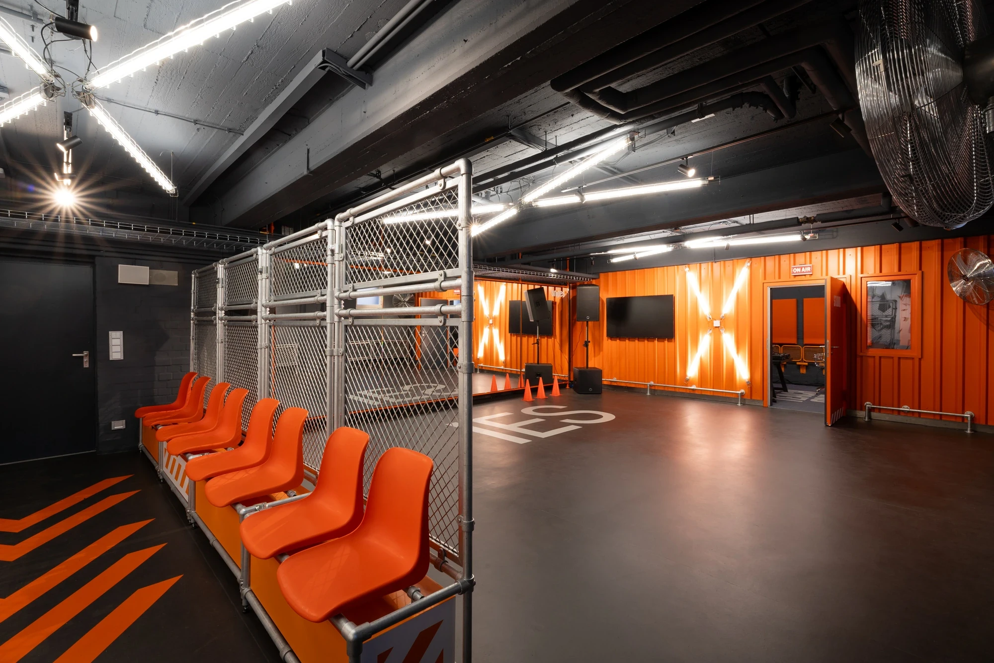 Training room with orange container walls, glowing X-shaped neon signage, bench seating behind a wire cage and screens for class instructions.
