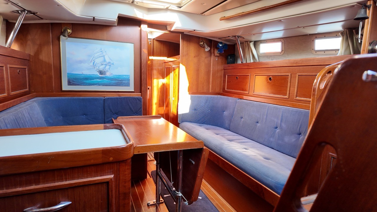 Contest 38 Ketch interior saloon — warm teak and mahogany joinery, blue buttoned settee berths port and starboard, fold-down saloon table, framed tall ship painting on bulkhead, oval portlights, and brass cabin lamp