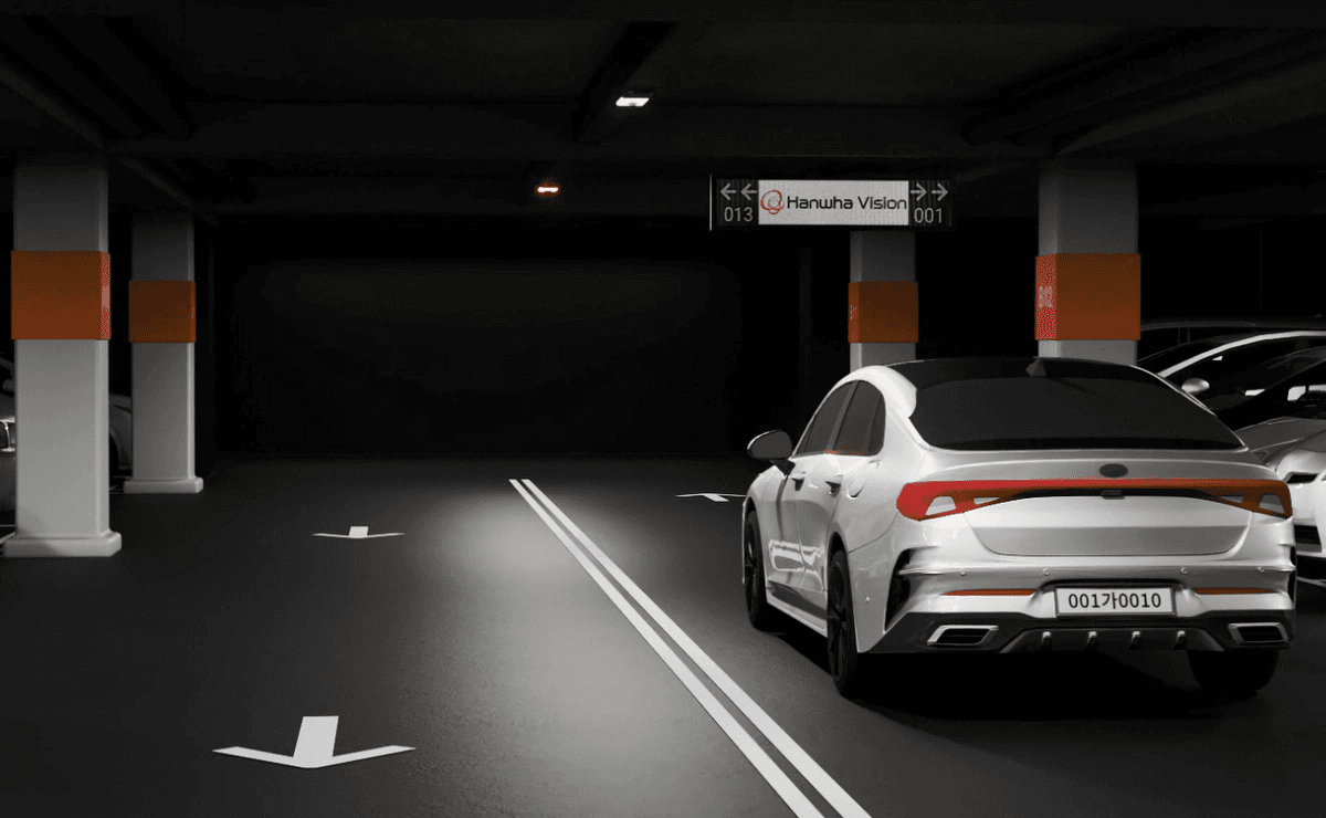 어두운 지하 주차장에서 흰색 차량이 이동 중이며, 천장에는 주차 가능 대수를 알려주는 디지털 현황판과 주차면의 점유 상태를 알려주는 녹색/적색 LED 표시등이 켜져 있는 모습입니다.