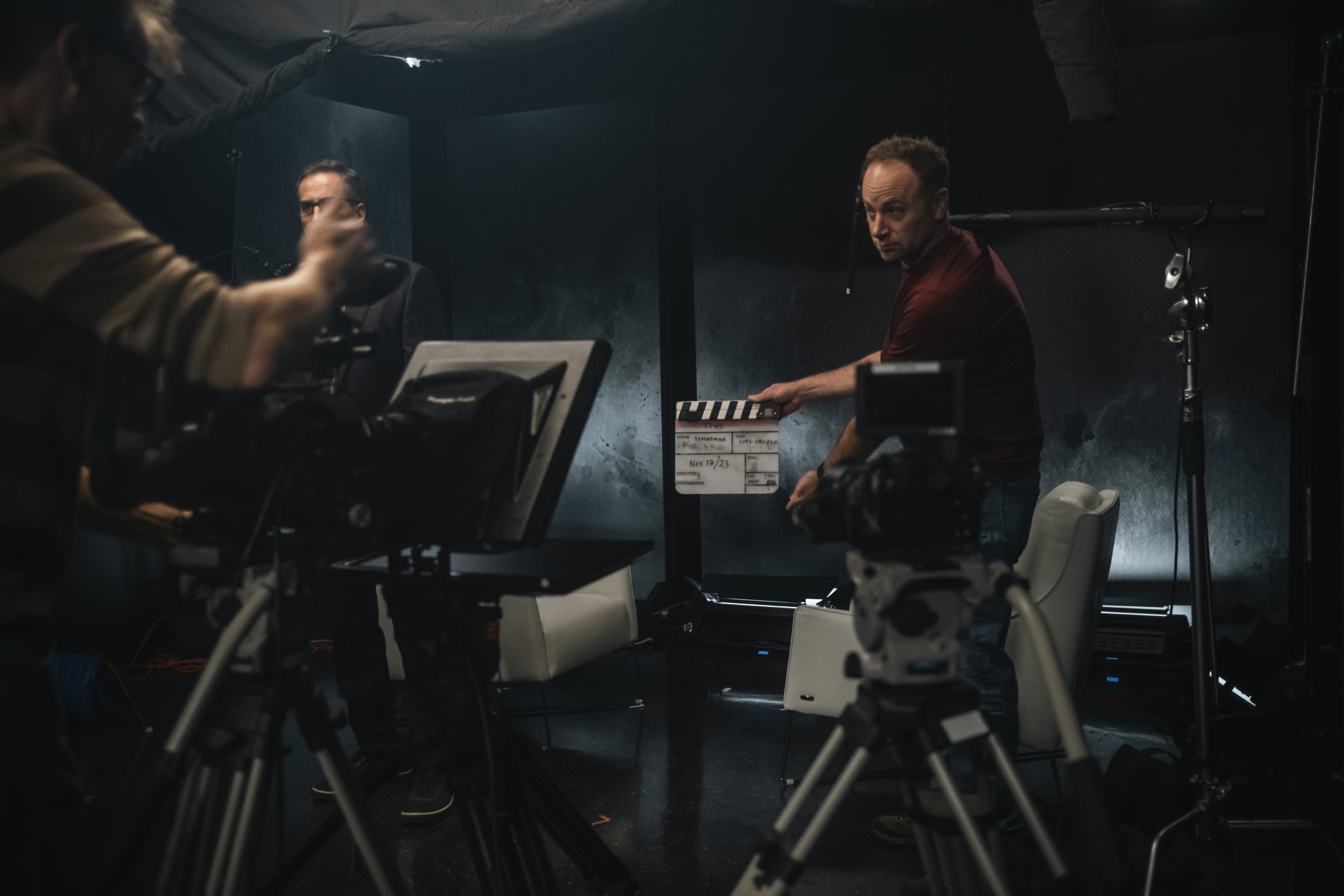 Two filmmakers operate a large camera on a dimly lit set. One wears headphones, both focused, suggesting a serious, creative atmosphere.
