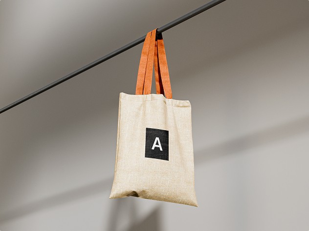 Beige fabric tote bag with orange handles hanging from a horizontal bar, featuring a black square logo with the letter “A” on the front.
