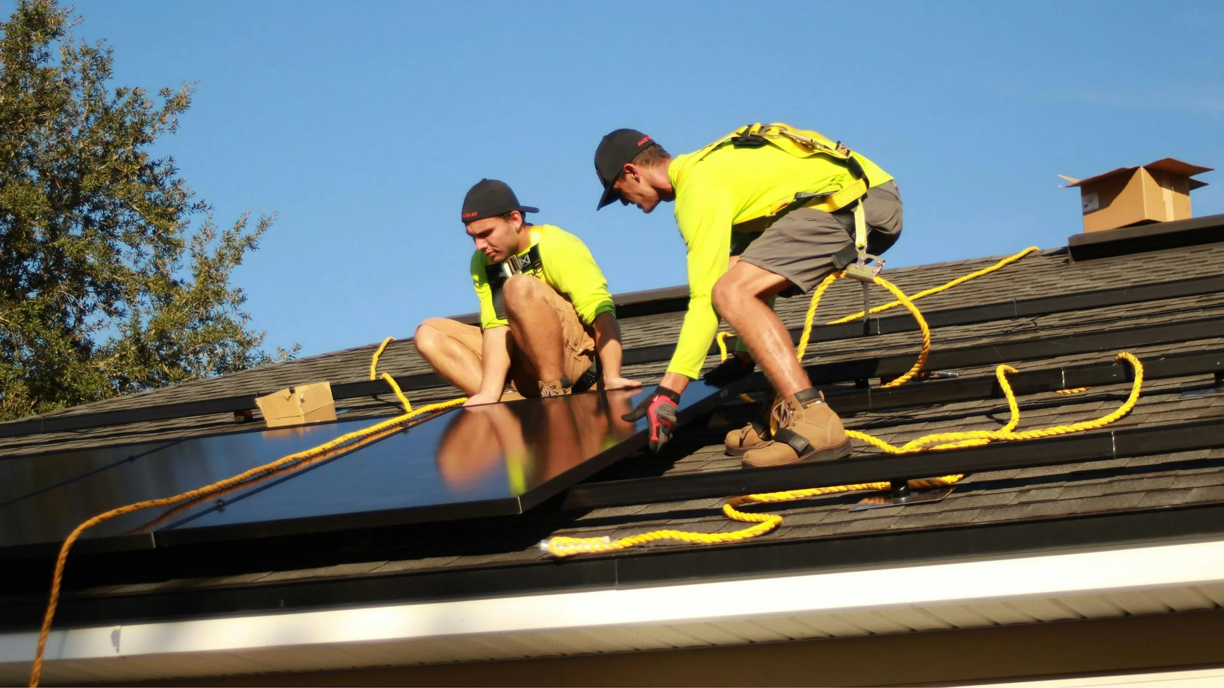 Two solar installers in safety harnesses mounting black panels on a residential roof