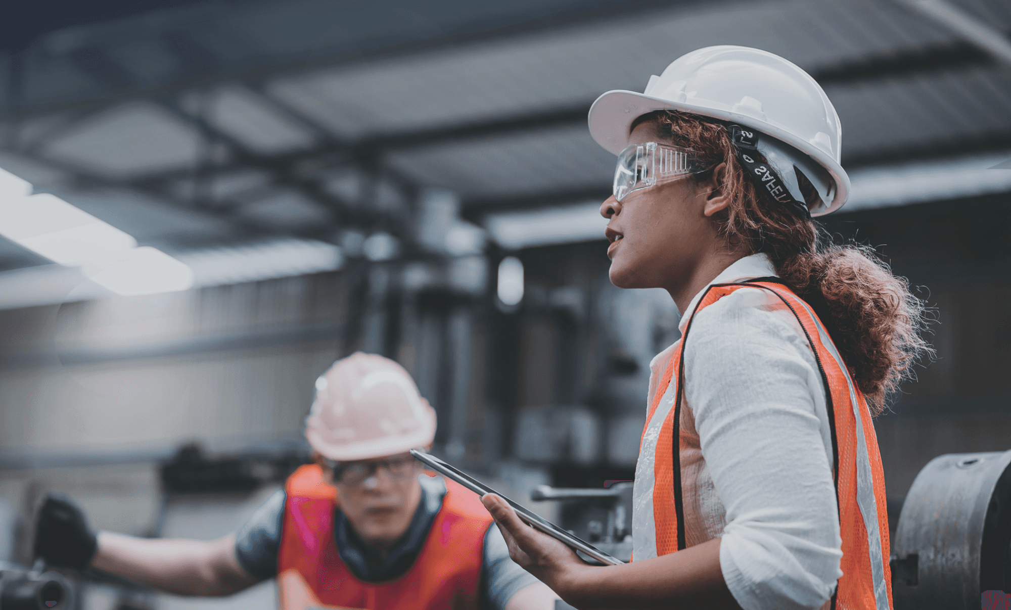 Two construction workers in hard hats and safety vests collaborate on a project in an industrial setting.