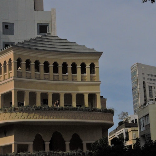 A beige building with arched windows and a terrace, surrounded by other buildings under a clear sky.