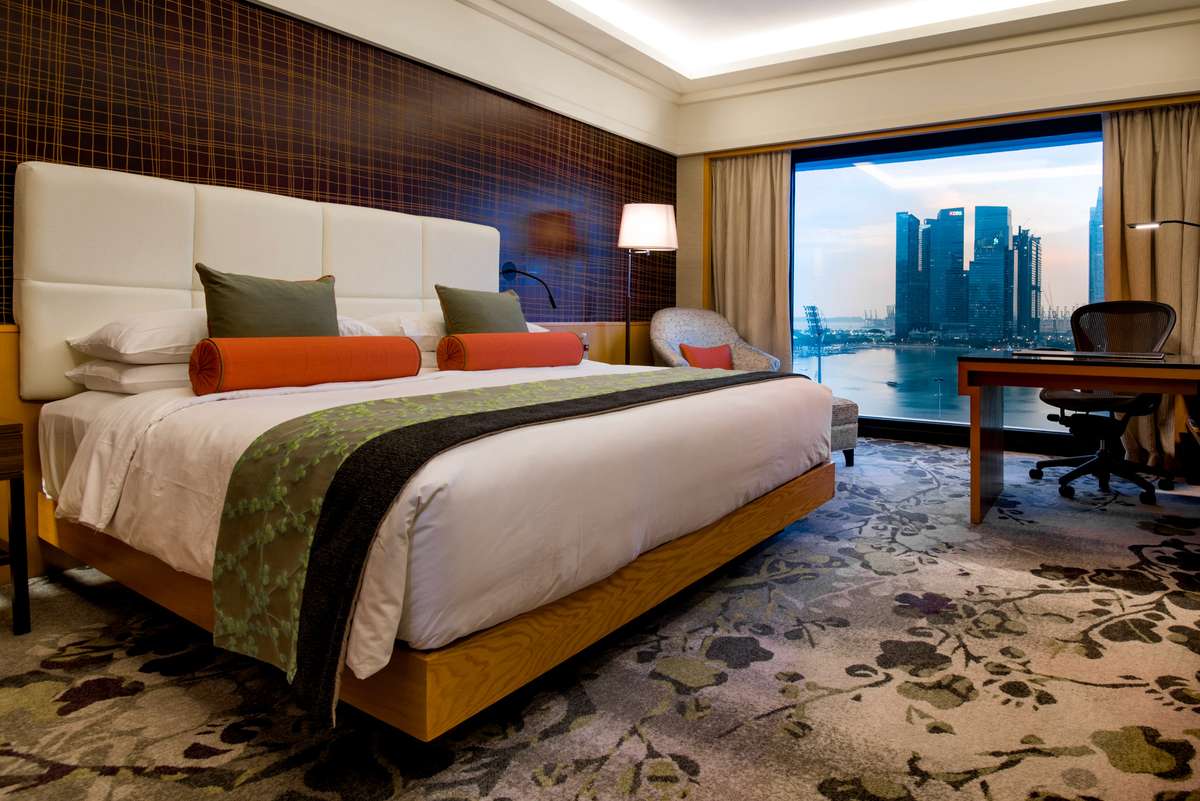 A modern hotel room with a large bed featuring orange and green pillows overlooks a city skyline across a body of water.