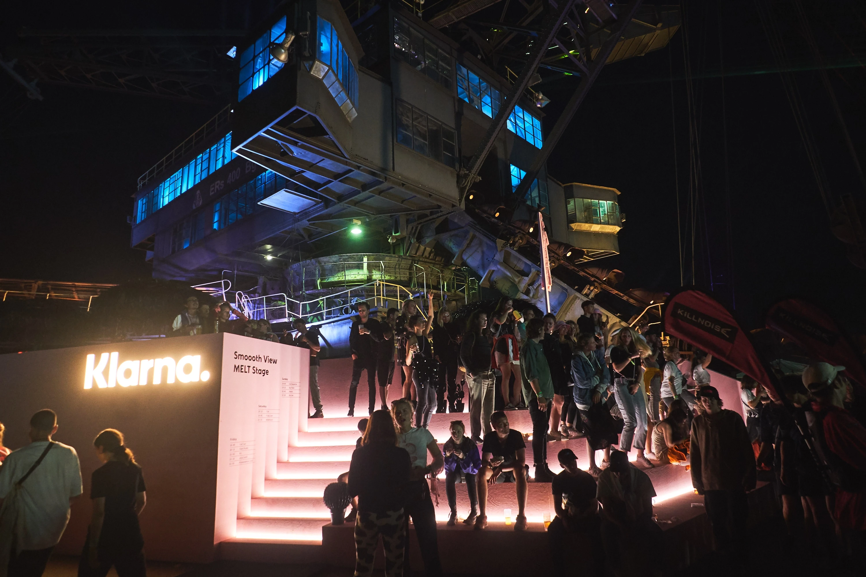 Festival visitors at night on the illuminated steps of the Klarna Smoooth View platform under the blue-lit excavator at Melt.