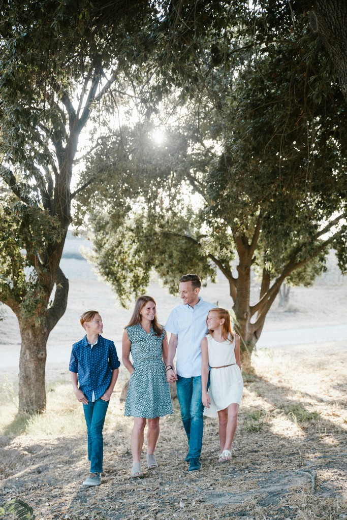 A happy family of four walks hand in hand under the shade of large trees in a sunlit park. The parents, along with their son and daughter, share smiles and conversation, capturing the essence of family togetherness and outdoor enjoyment on a beautiful day.