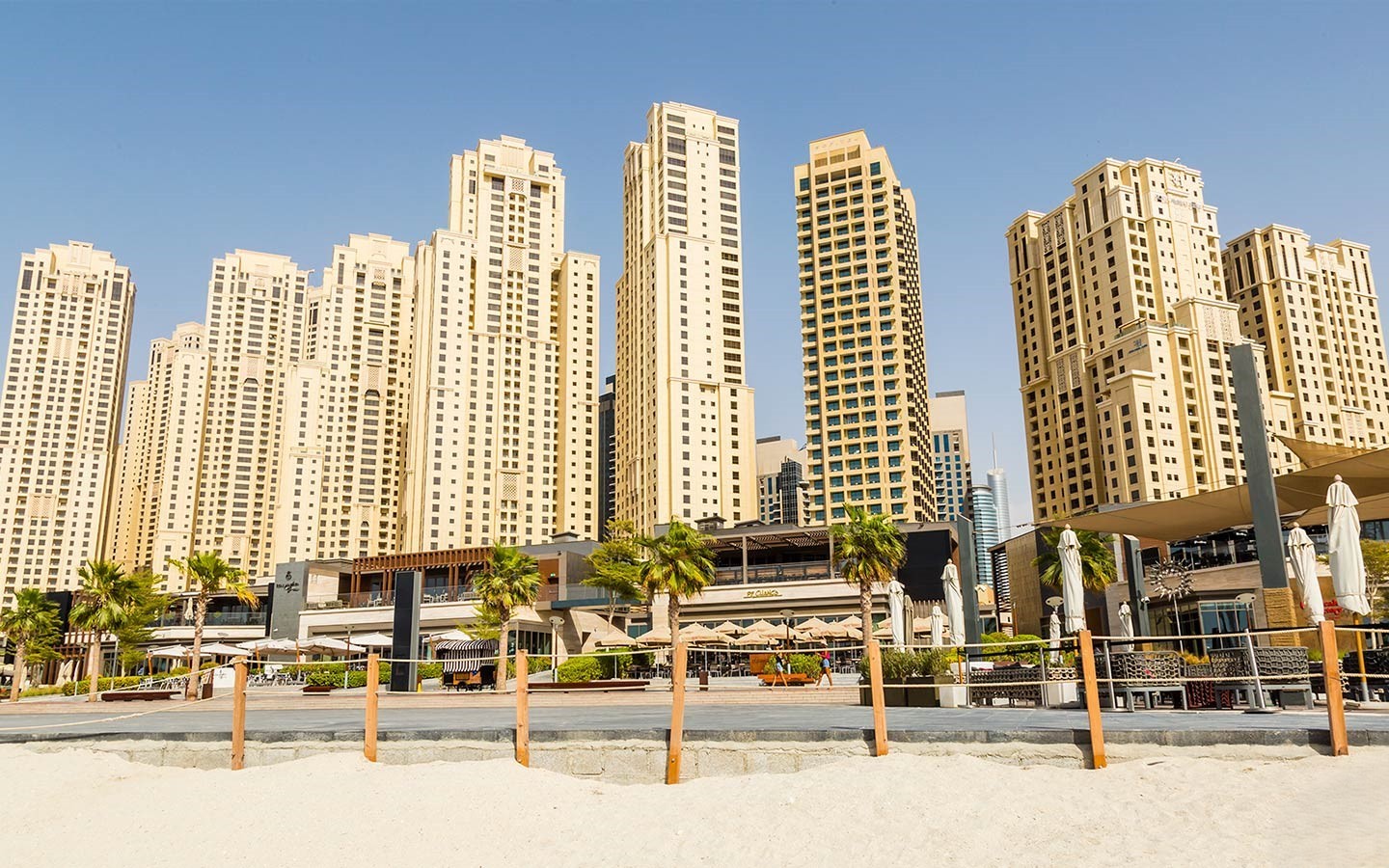 View of Jumeirah Beach Residence towers rising behind beachfront cafés and palm trees under a clear blue sky.