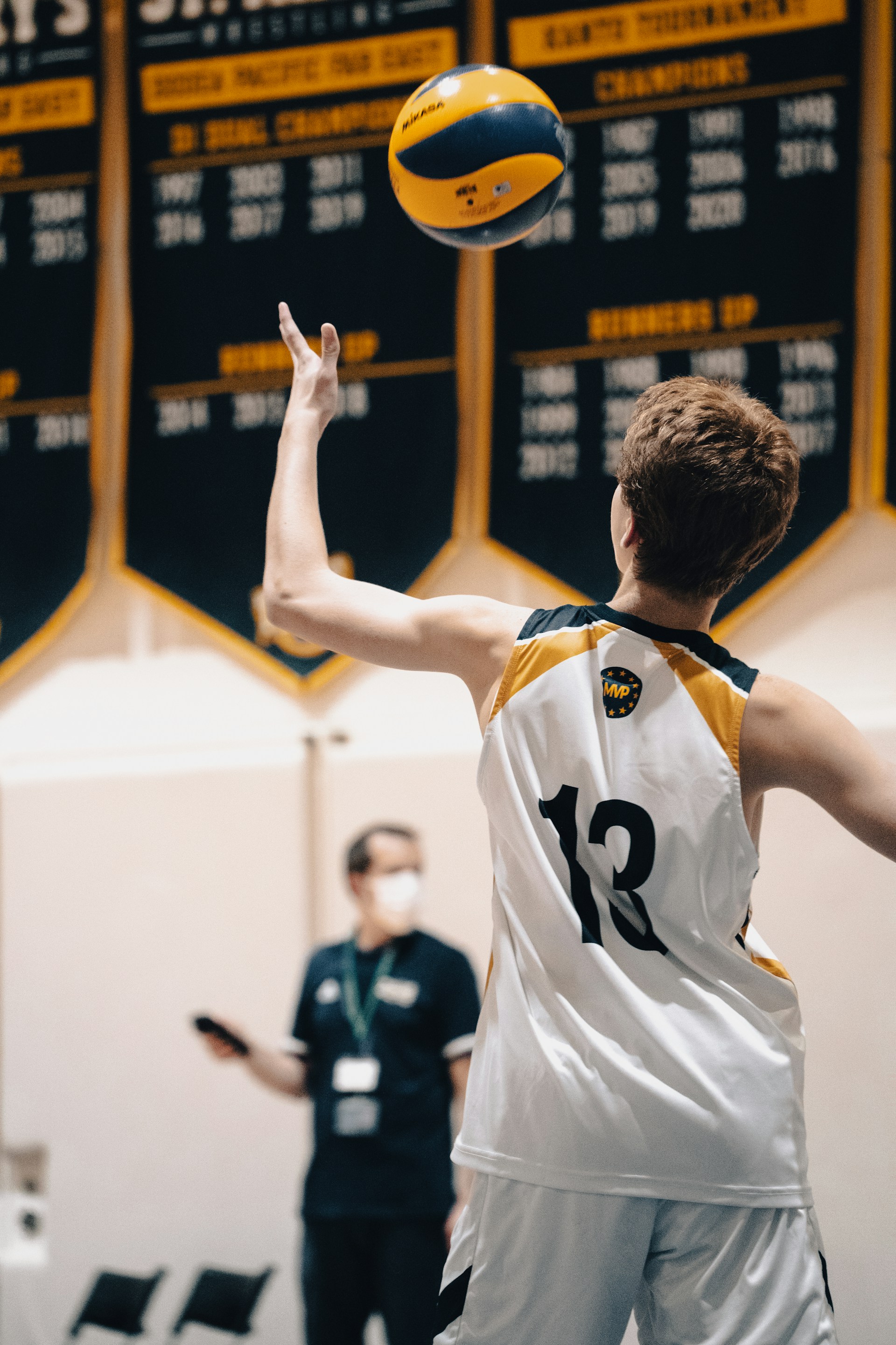 Rear view of a volleyball player in a white jersey (number 13) tossing the ball up to serve.