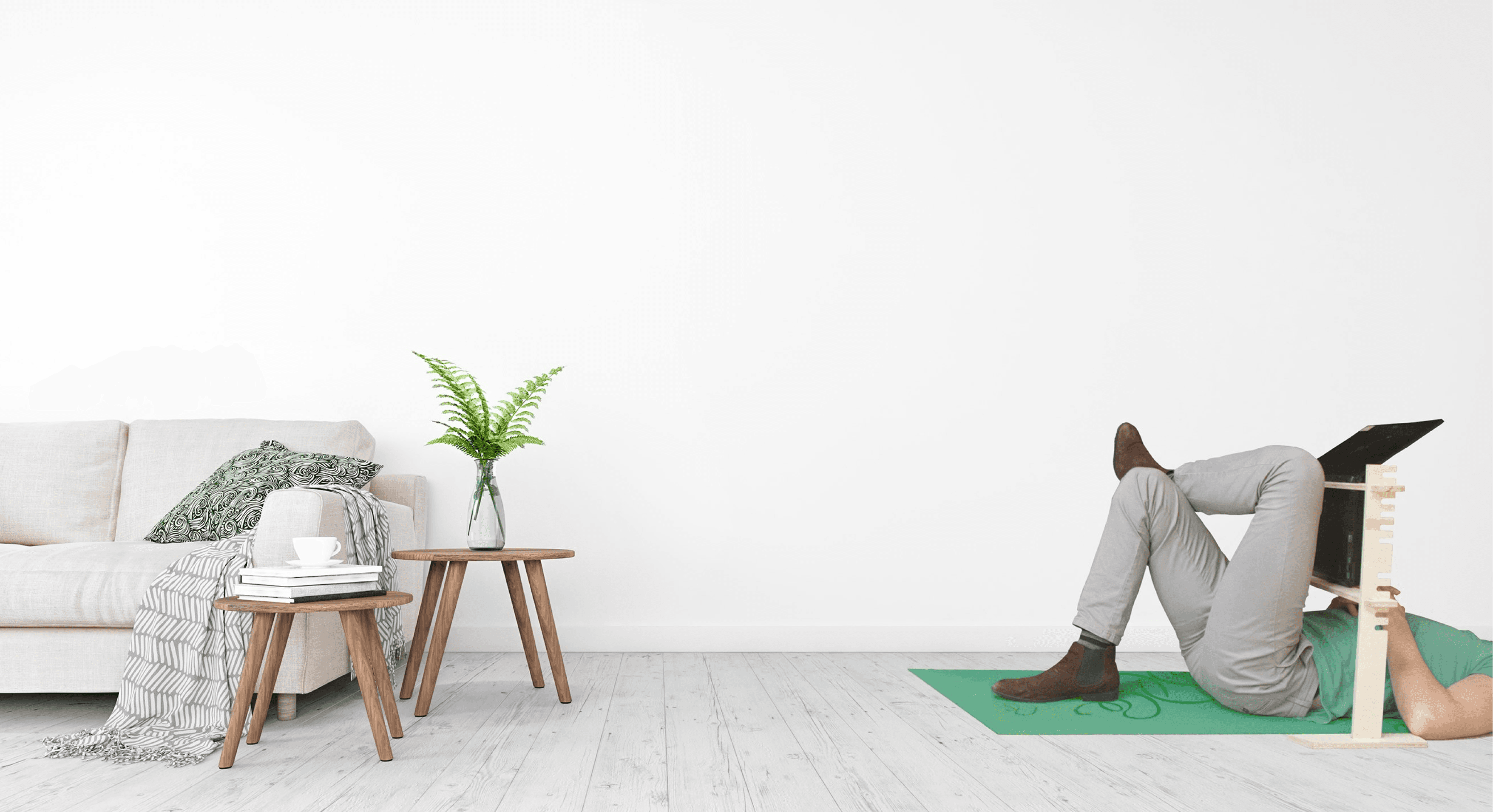 A person using a lie down laptop stand in the right of the image with a sofa and small tables and plants to the left.. They are wearing a green tshirt and lying on a green yoga mat. The person, who's head is out of shot, is wearing grey pants. The wall and floor are also grey.