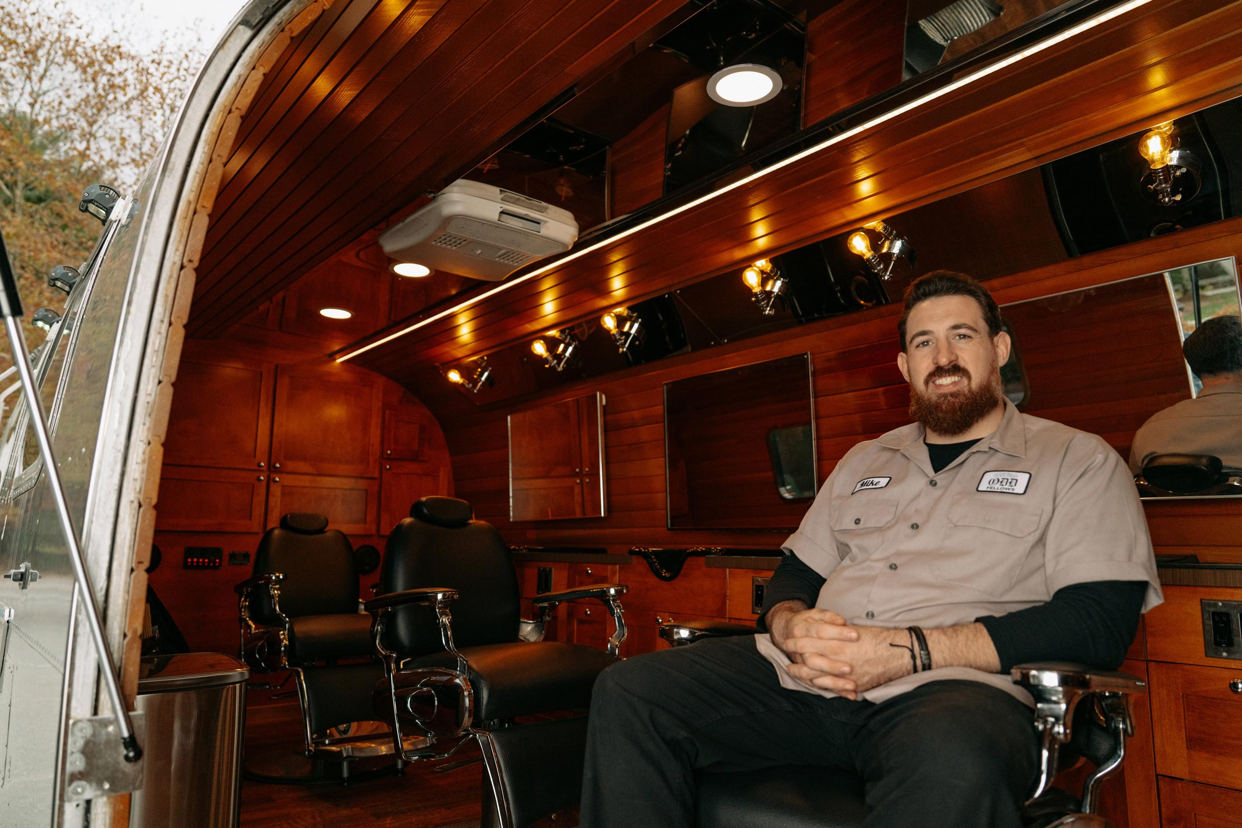 A man with a full beard and a tan work shirt is sitting on a cushioned bench inside a customized mobile barbershop housed in a silver Airstream-style trailer. The interior features rich, dark wood paneling, recessed lighting, and two black leather barber chairs with vanity mirrors. The man is looking directly at the camera with a slight smile.