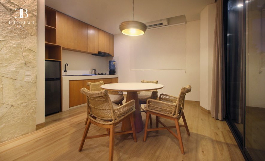 A warm and modern kitchen and dining area at Echo Beach Suite in Canggu, Bali, featuring wooden cabinetry, a compact kitchenette, woven rattan dining chairs, and soft lighting, ideal for a relaxed stay near Echo Beach.