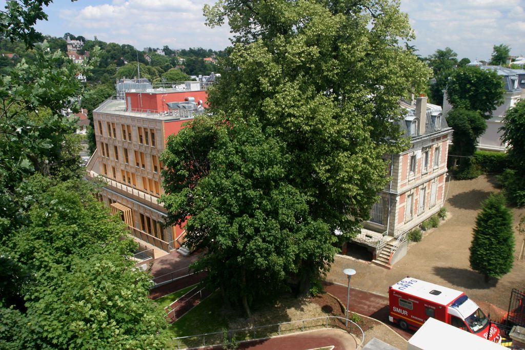 Multi-story residential building with red brick facade, surrounded by lush green trees and vegetation, internal courtyard with green lawn visible