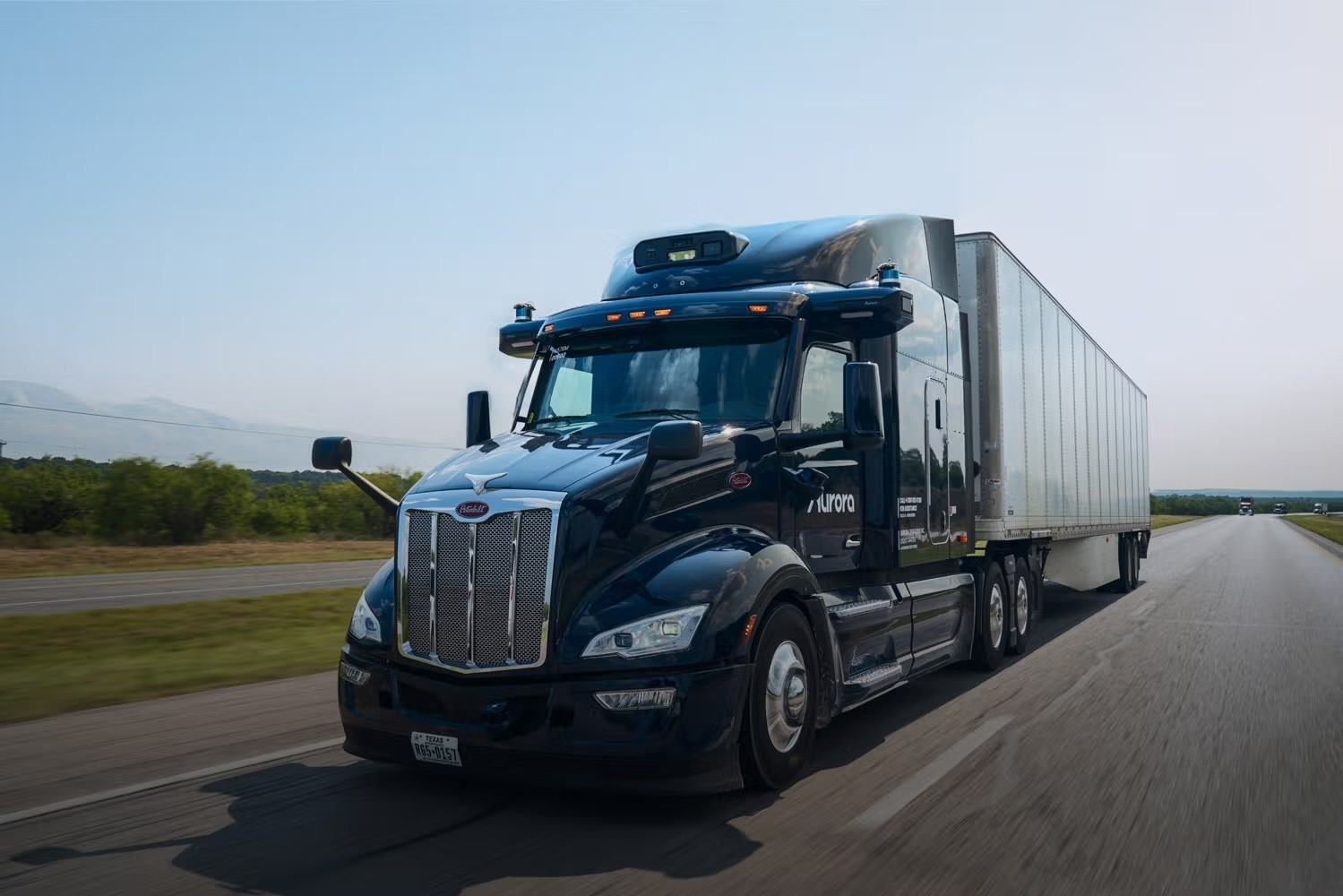 Close-up front view of an Aurora self-driving semi-truck on a highway. The truck features a distinctive sensor array mounted on top of the cab, including lidar and camera systems for autonomous navigation. The truck has a modern aerodynamic design with a white and blue color scheme. The Aurora branding is visible on the front of the vehicle.