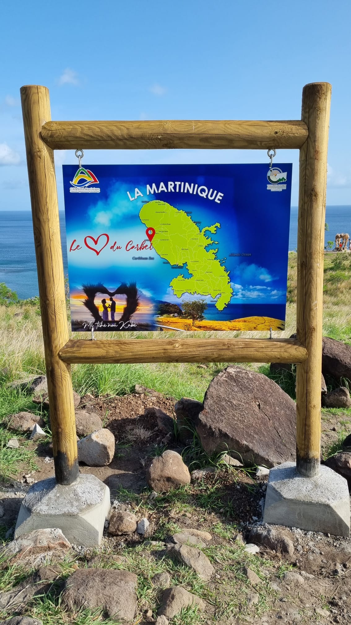 Wooden-framed sign displaying a map of Martinique under blue skies. The text reads, "La Martinique" with a heart symbol. Ocean visible in the background.