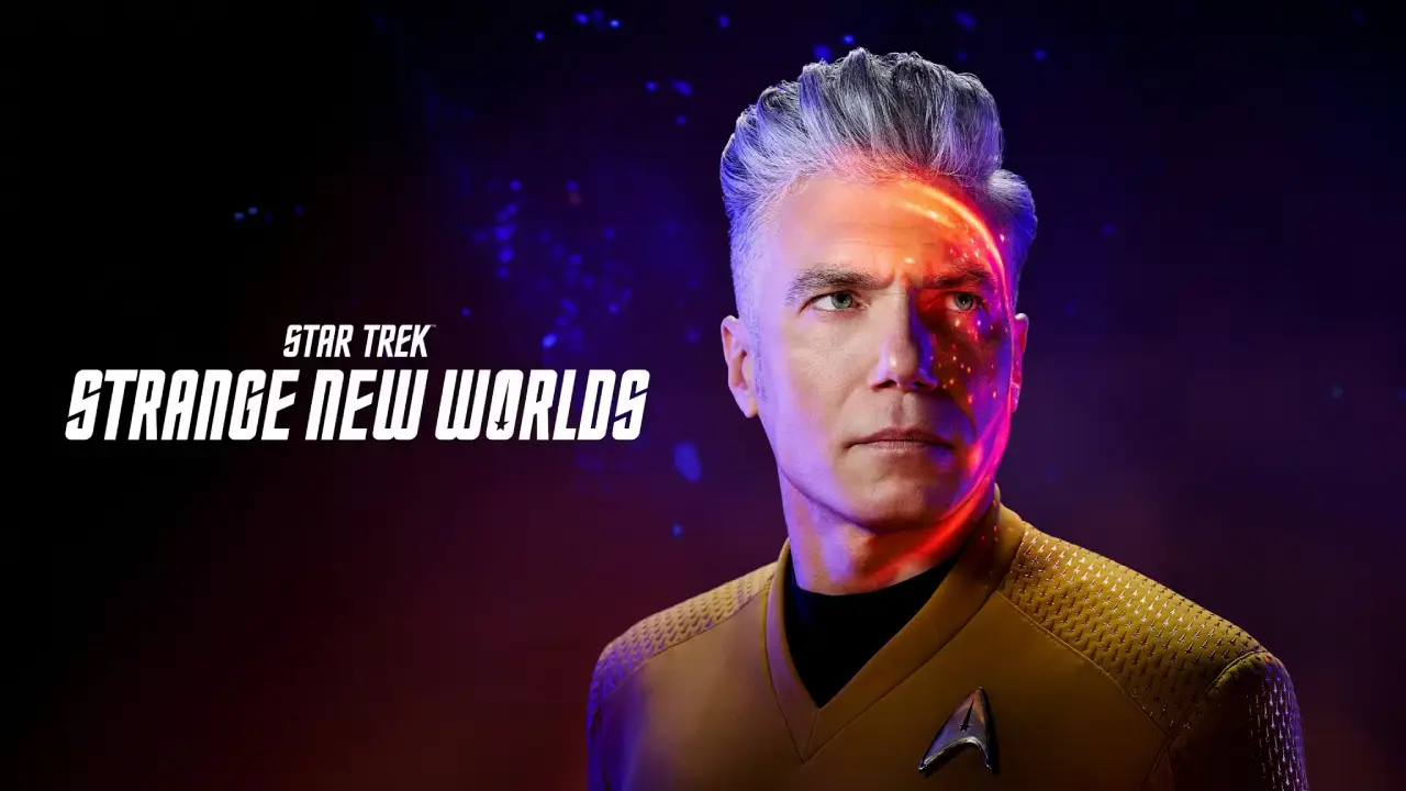 A man in a futuristic yellow uniform is illuminated by dramatic lighting, with a vibrant orange glow on their face and a galaxy backdrop featuring text that reads "Star Trek Strange New Worlds."