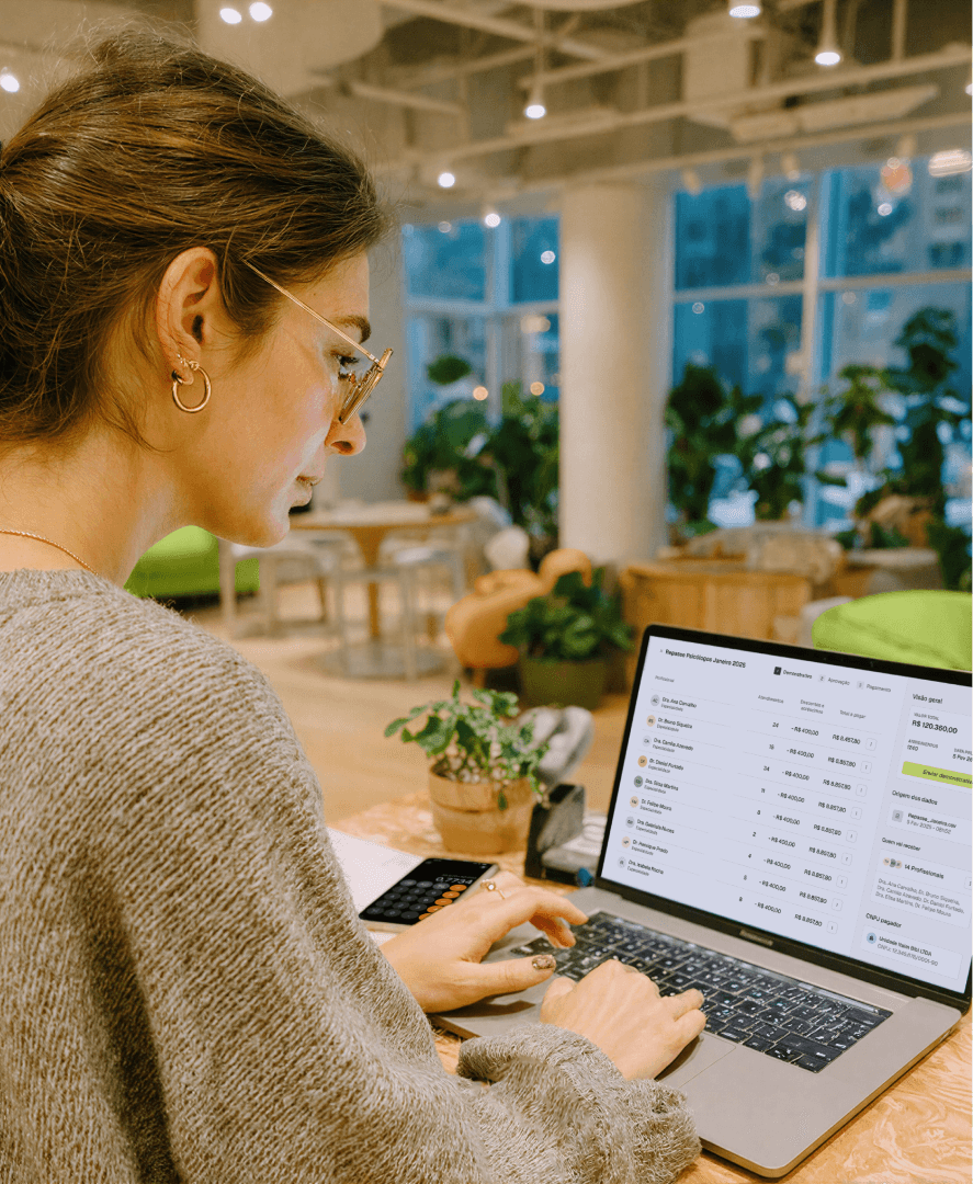 Mulher de óculos trabalhando em um notebook em uma clínica, com o painel financeiro da Tivita aberto na tela para conciliação bancária em tempo real.
