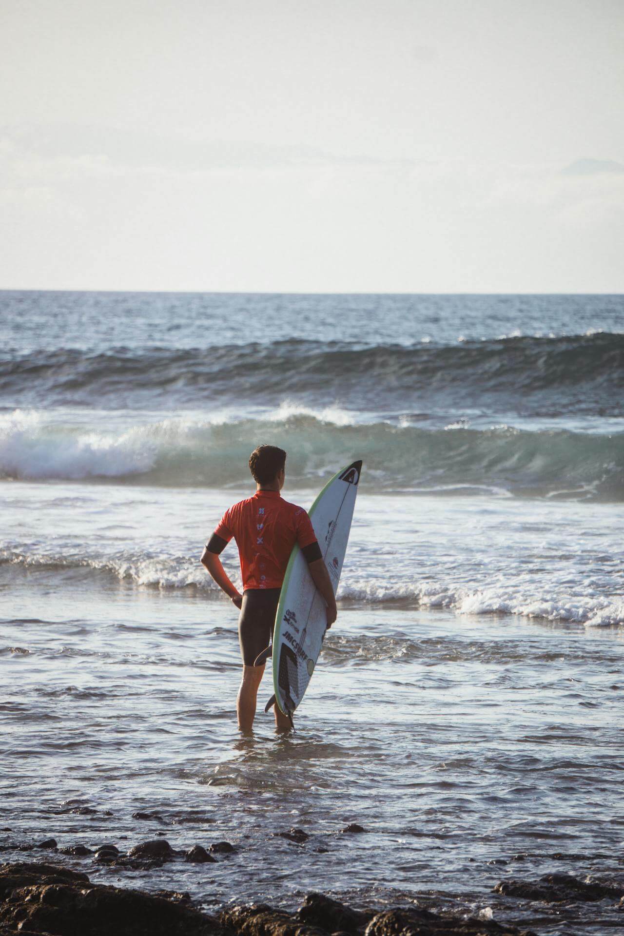 A surfer stands at the edge of the ocean, watching the waves before paddling out, surrounded by golden light and quiet surf.