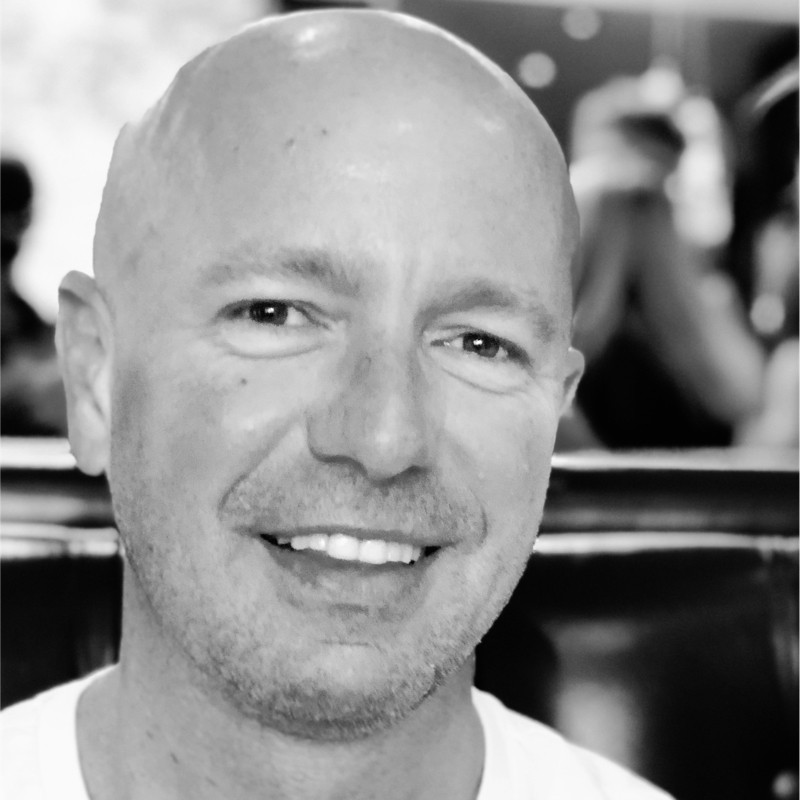 A smiling man with short hair stands with arms crossed, wearing a dark shirt against a neutral background.
