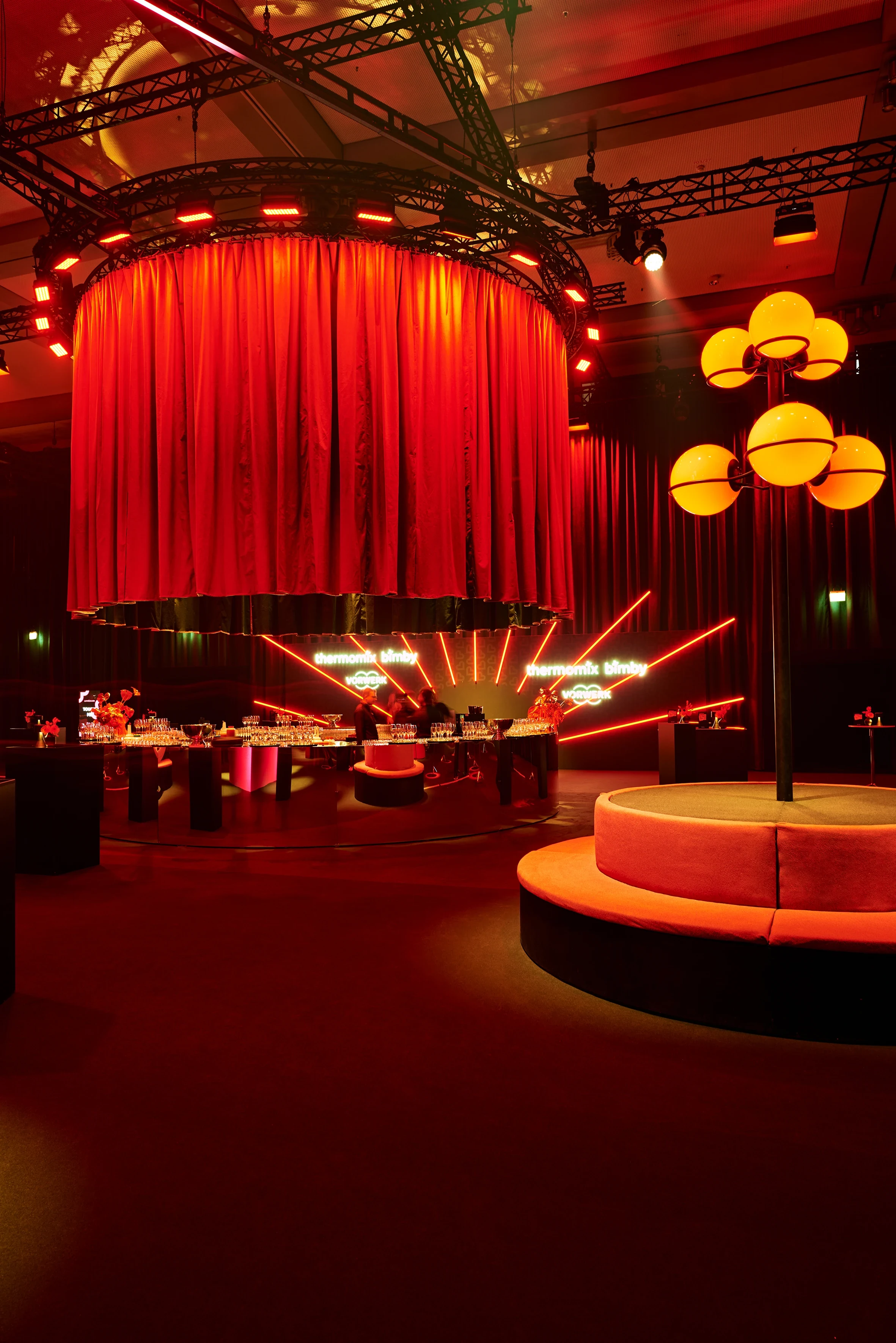 Red event hall with giant curtain cylinder, lantern sculpture and round velvet bench.