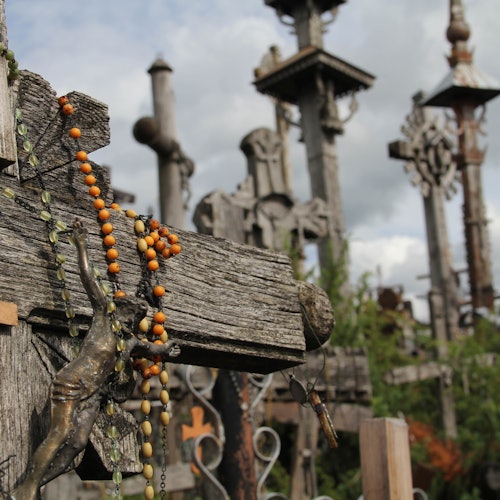 Numerous weathered wooden crosses and rosary beads are seen in an outdoor area under a cloudy sky.