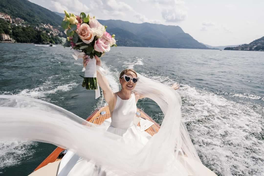 Bride on a boat during a destination wedding, dress flowing in the wind with mountains and lake in the background
