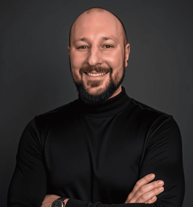A smiling man with curly hair is sitting at a desk, looking at the camera, with a laptop and a lamp in the background.