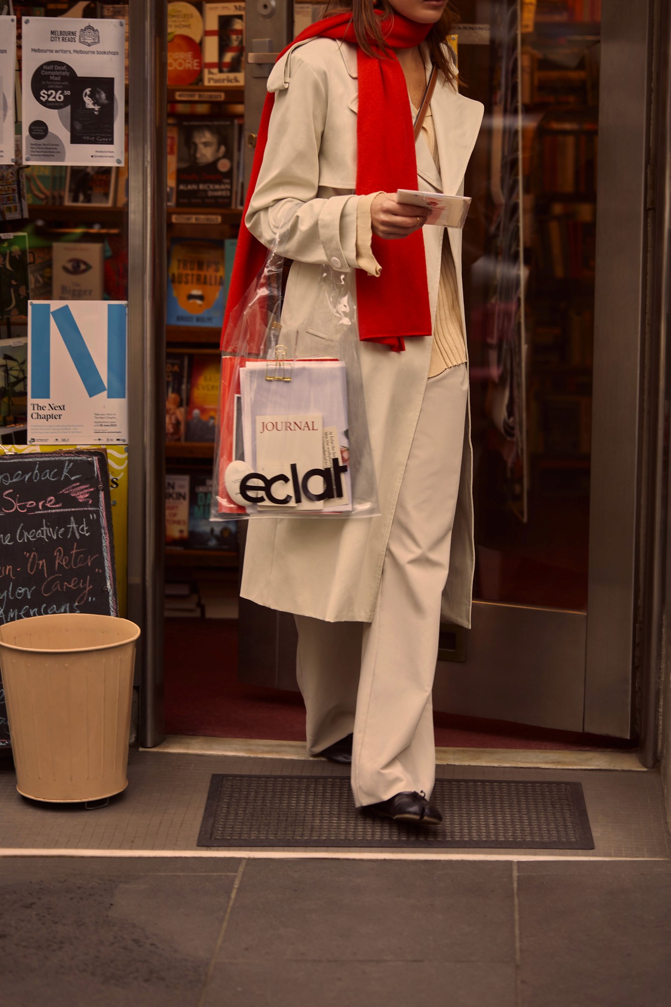 Person carrying a clear tote bag holding an eclat journal and papers while leaving a bookstore, with a red scarf and chalkboard sign visible
