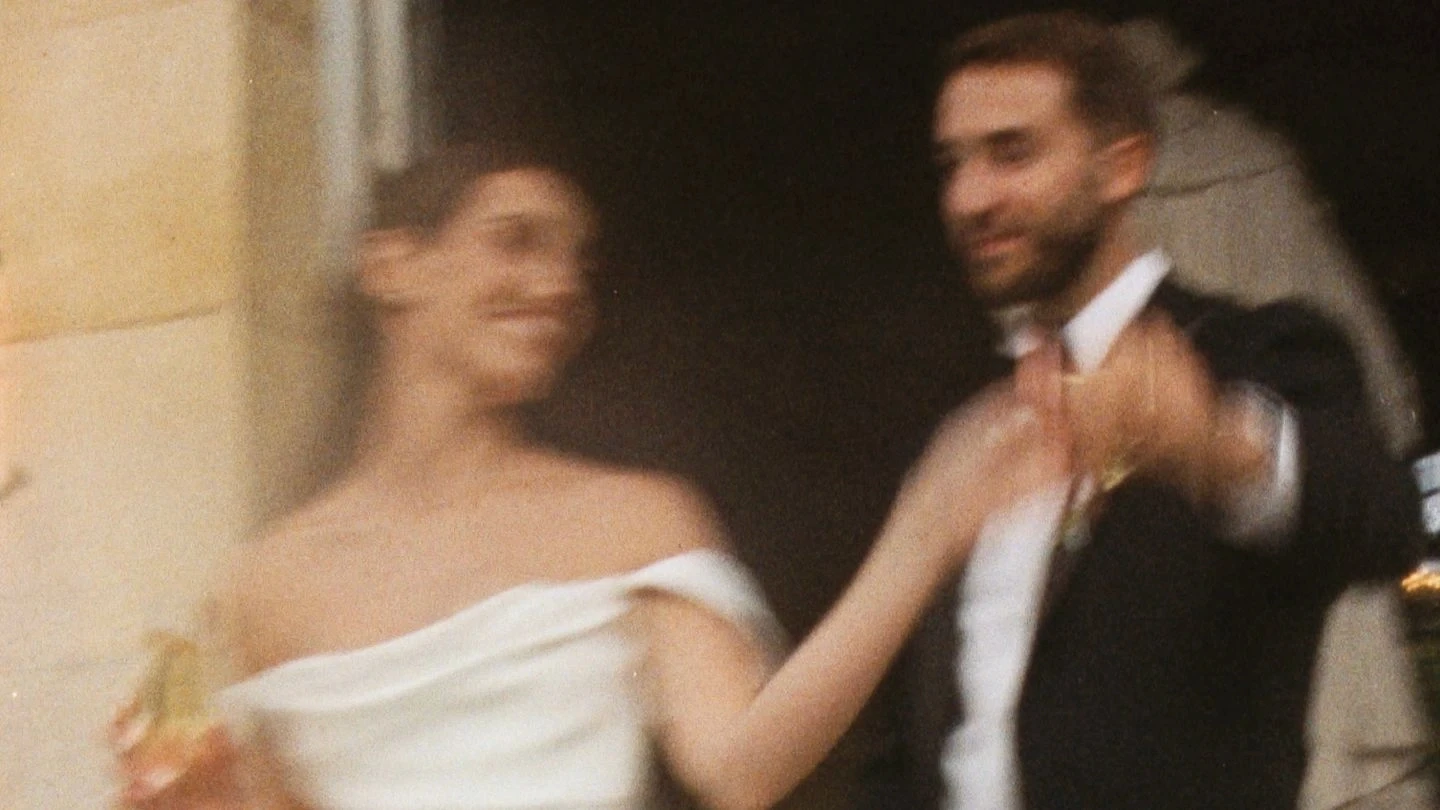 A blurred image of a couple in formal attire, possibly at a wedding, with the woman in an off-shoulder dress holding a champagne glass and the man in a dark suit extending his hand.