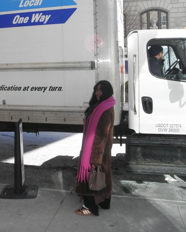 Person wearing a blue puffer jacket and red backpack standing in front of a light blue building on a city street.