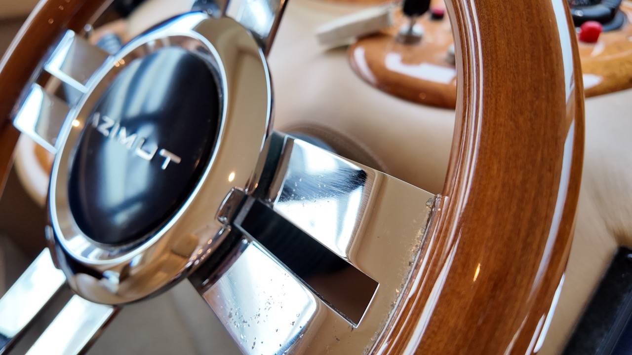 Close-up detail of the Azimut 68 Evolution branded wood-rim helm wheel with polished stainless steel spokes and chrome Azimut centre cap, showing the burl walnut dashboard, joystick docking system, and engine throttle controls in the background
