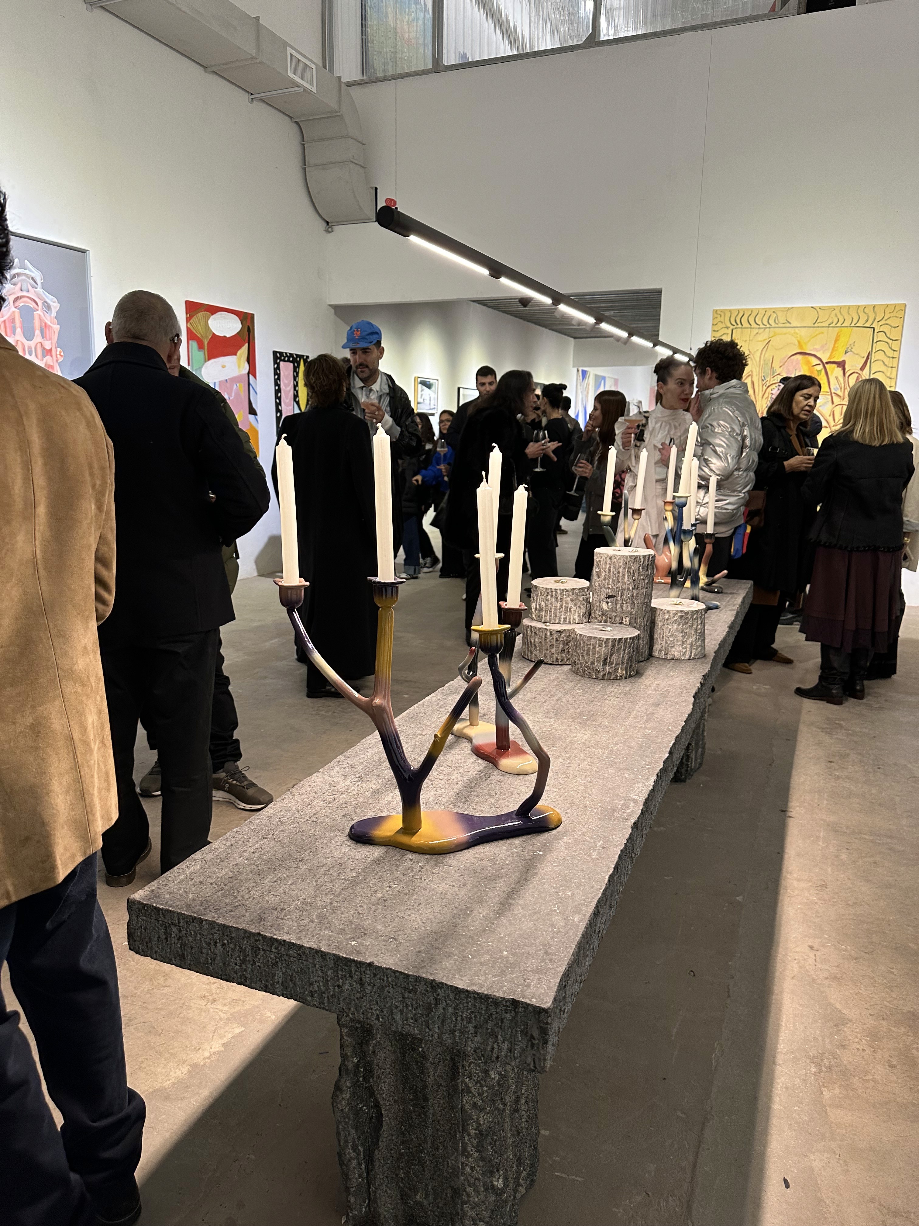 En la foto se observa una mesa larga con candelabros de diseño único y contemporáneo. Personas en conversación a su alrededor, lo que sugiere un evento social o una apertura. El fondo muestra más obras de arte, ofreciendo un vistazo a la variedad de estilos presentes en la exposición.