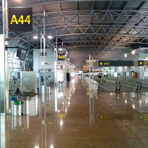 Prázdné letištní terminál s vysokým stropem, ukazateli bran (A44, A46), pohyblivými chodníky a lesklou podlahou.