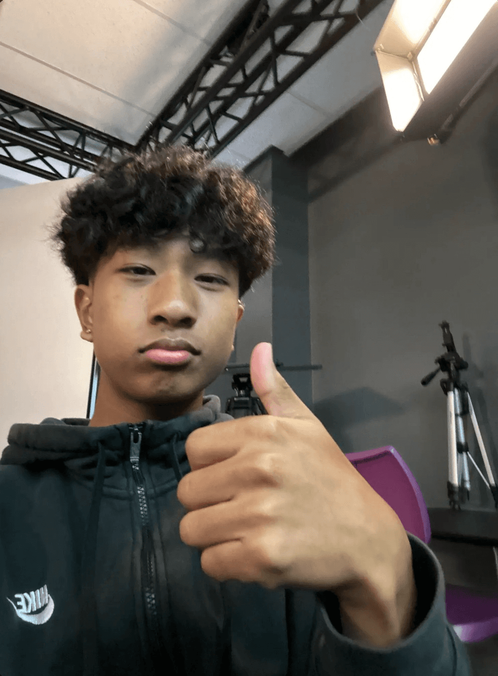 A selfie of Steven Fang wearing a black Nike zip-up hoodie, giving a thumbs up to the camera. He is indoors in a production studio setting, with a lighting grid on the ceiling, a large studio light, and a tripod visible in the gray background.