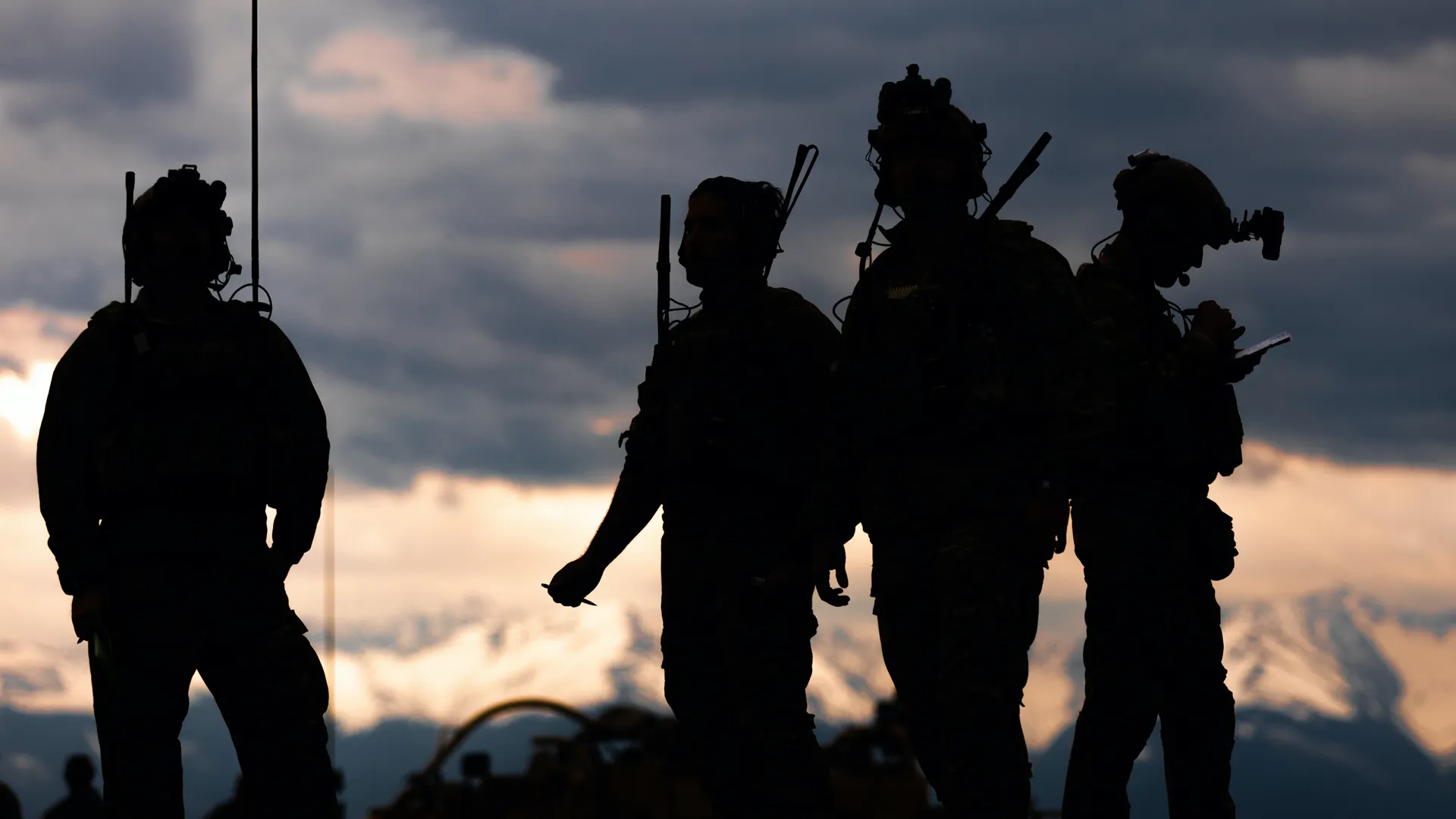 Silhouettes of four soldiers with tactical and communication gear against a dramatic dusk sky.