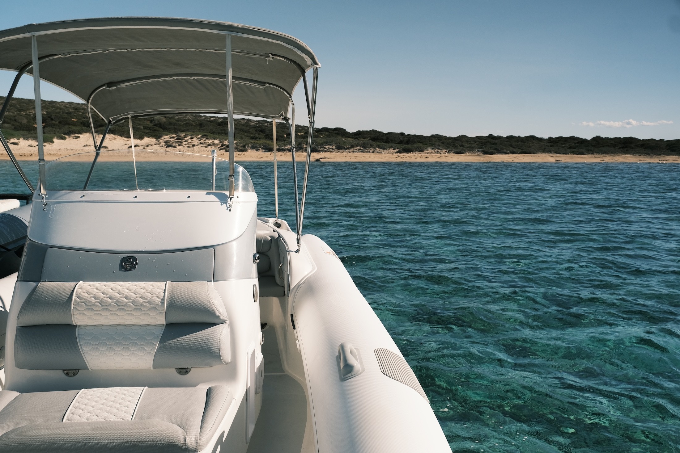 White motor yacht with sun canopy anchored in crystal clear turquoise waters near a sandy beach with vegetation in the Cyclades.