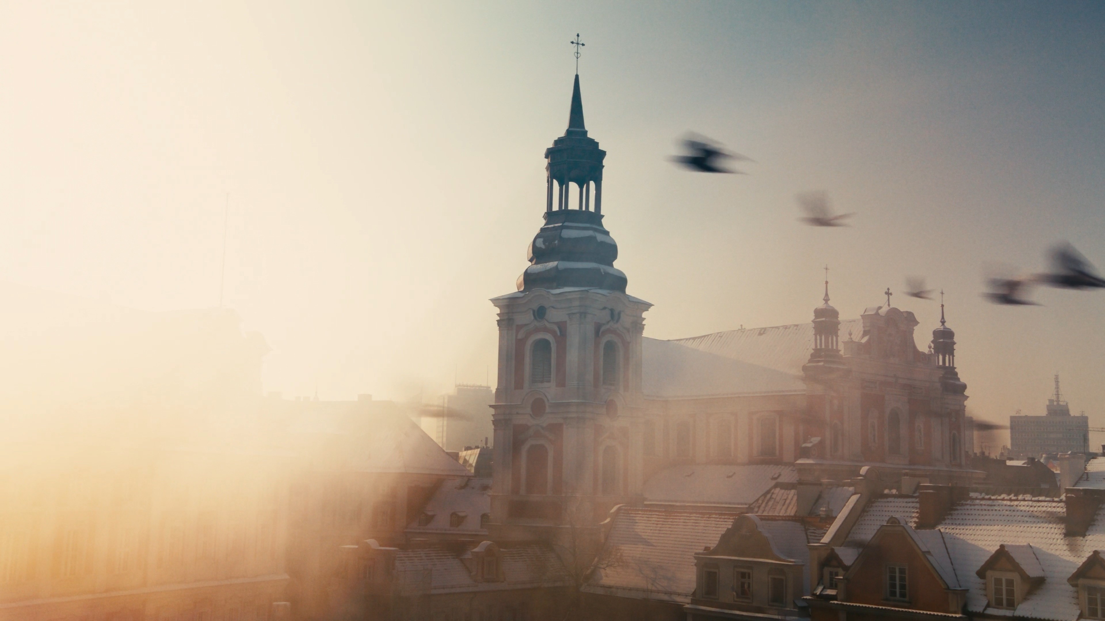 Establishing shot of a church tower and city rooftops shrouded in mist and sunlight.