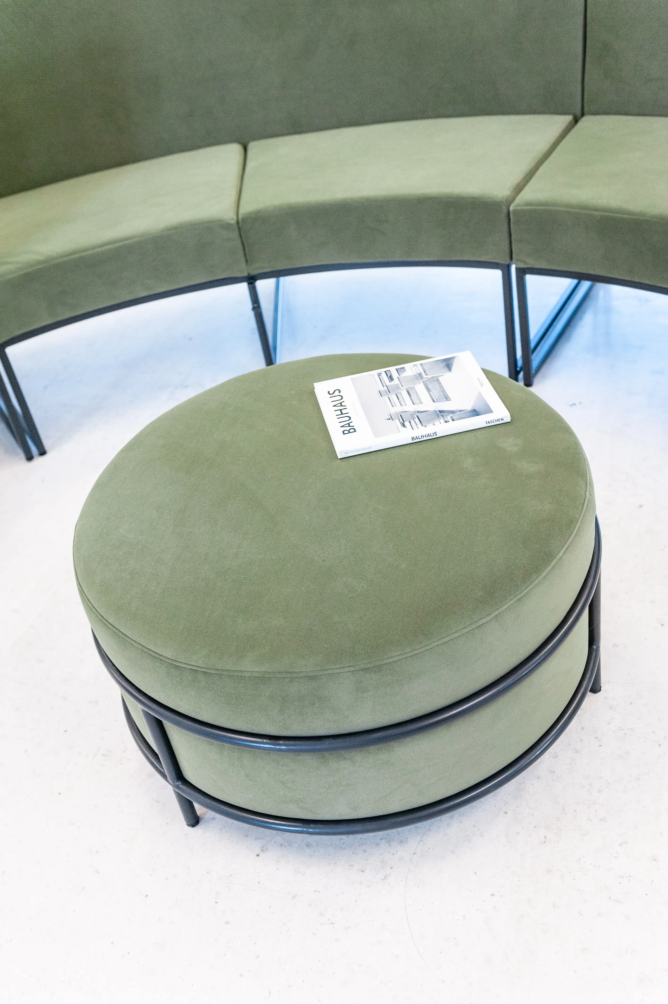 Close-up of a round sage-green velvet pouf in a black steel frame with a Bauhaus magazine on top, next to a curved green upholstered bench at LA RED.