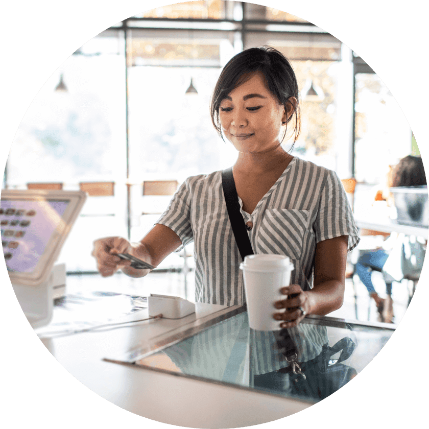 A woman in a striped shirt standing at a cafe counter, using a card to pay while holding a coffee cup. The interior features large windows and soft lighting.