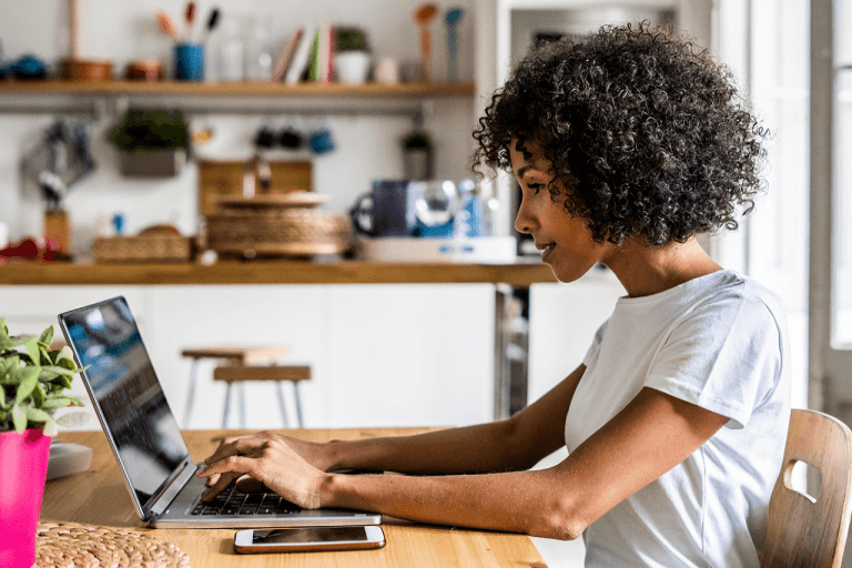 Een vrouw met krullend haar tikt op een laptop in een knusse werkruimte met planken en planten op de achtergrond.