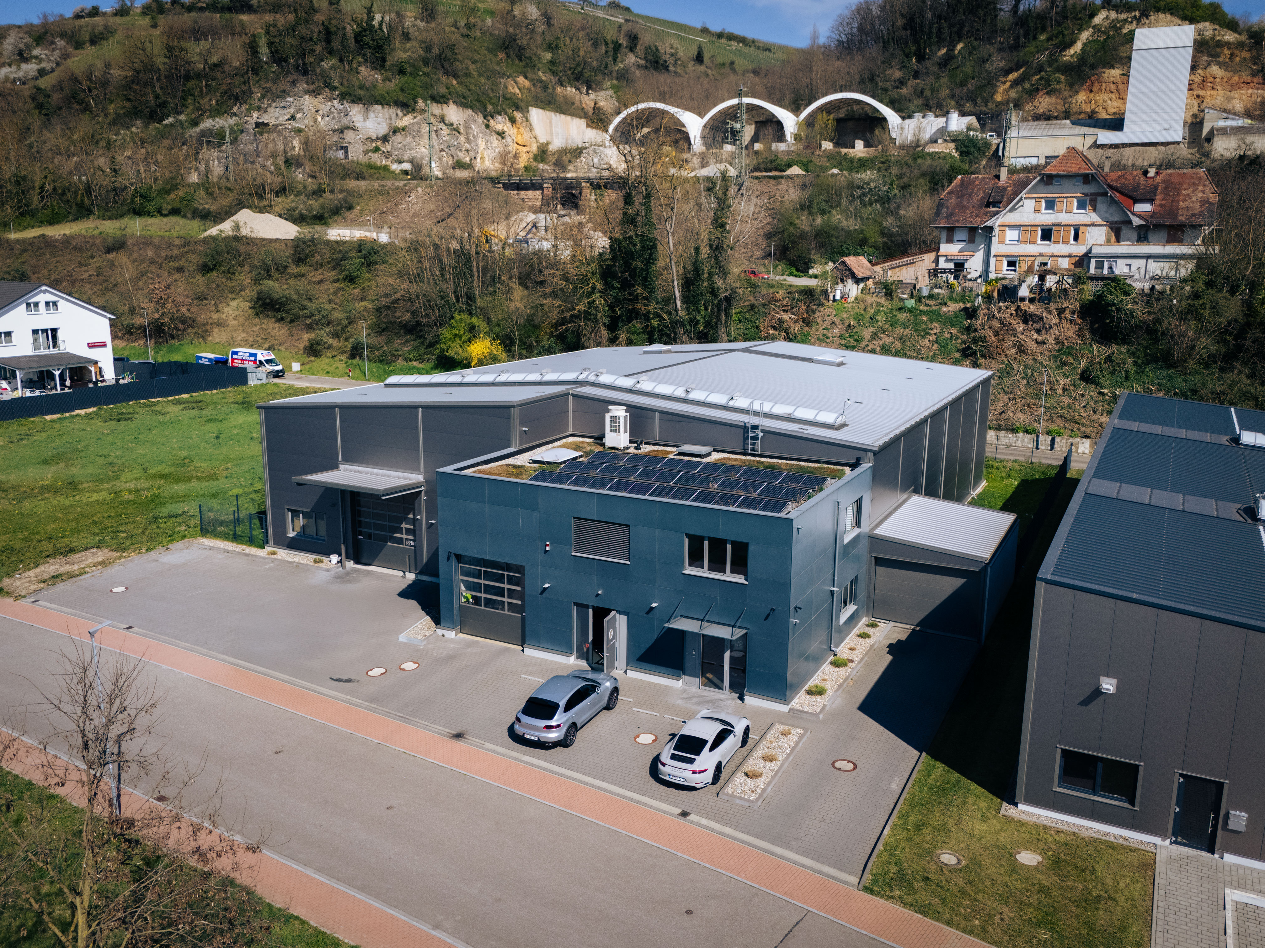 Luftaufnahme eines modernen Firmengebäudes mit Solaranlage auf dem Dach und zwei geparkten Fahrzeugen, gelegen in einer grünen Umgebung mit Felsen im Hintergrund