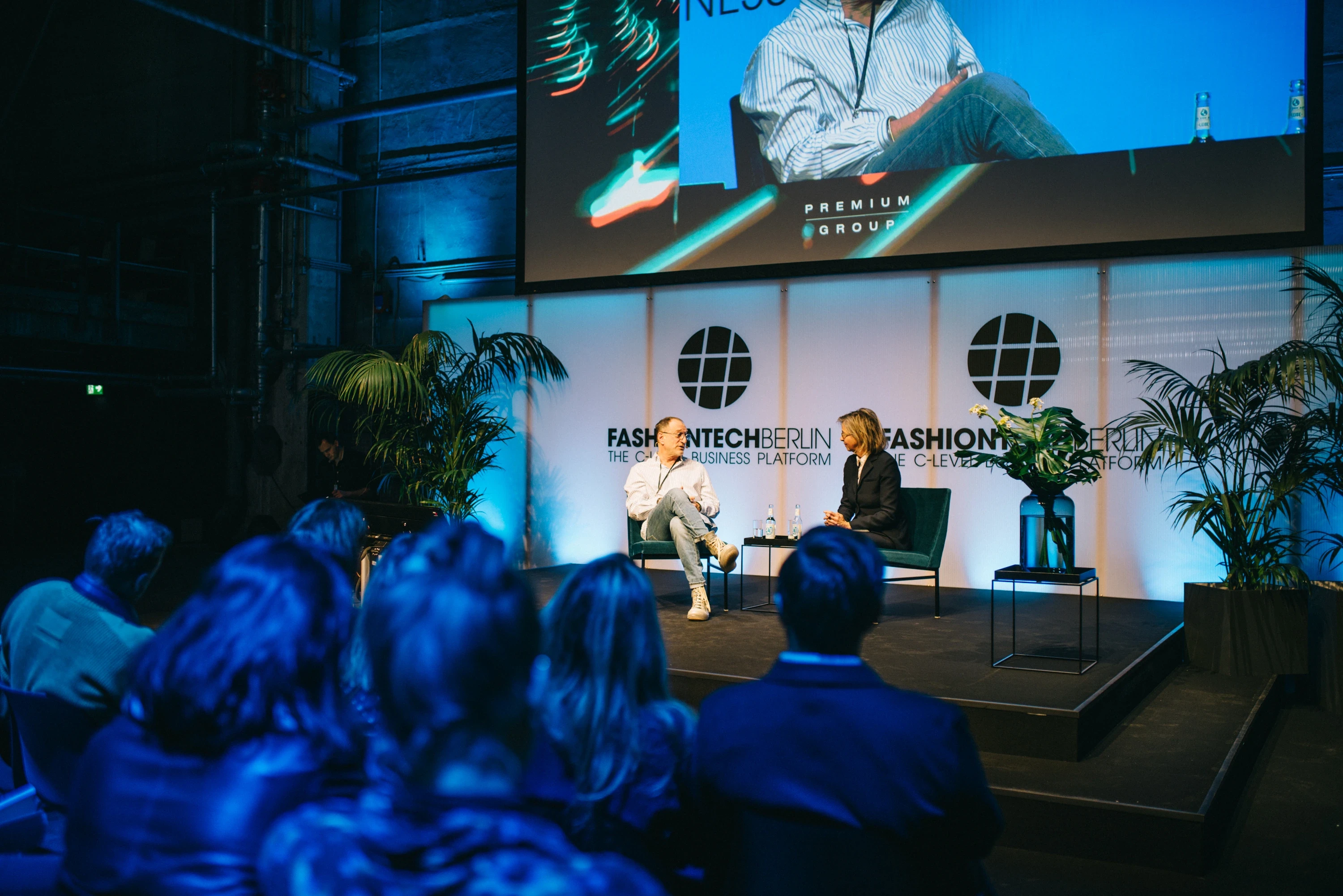 Panel talk on the FASHIONTECH Berlin stage with two speakers in green armchairs in front of backlit logo walls and palm plants.