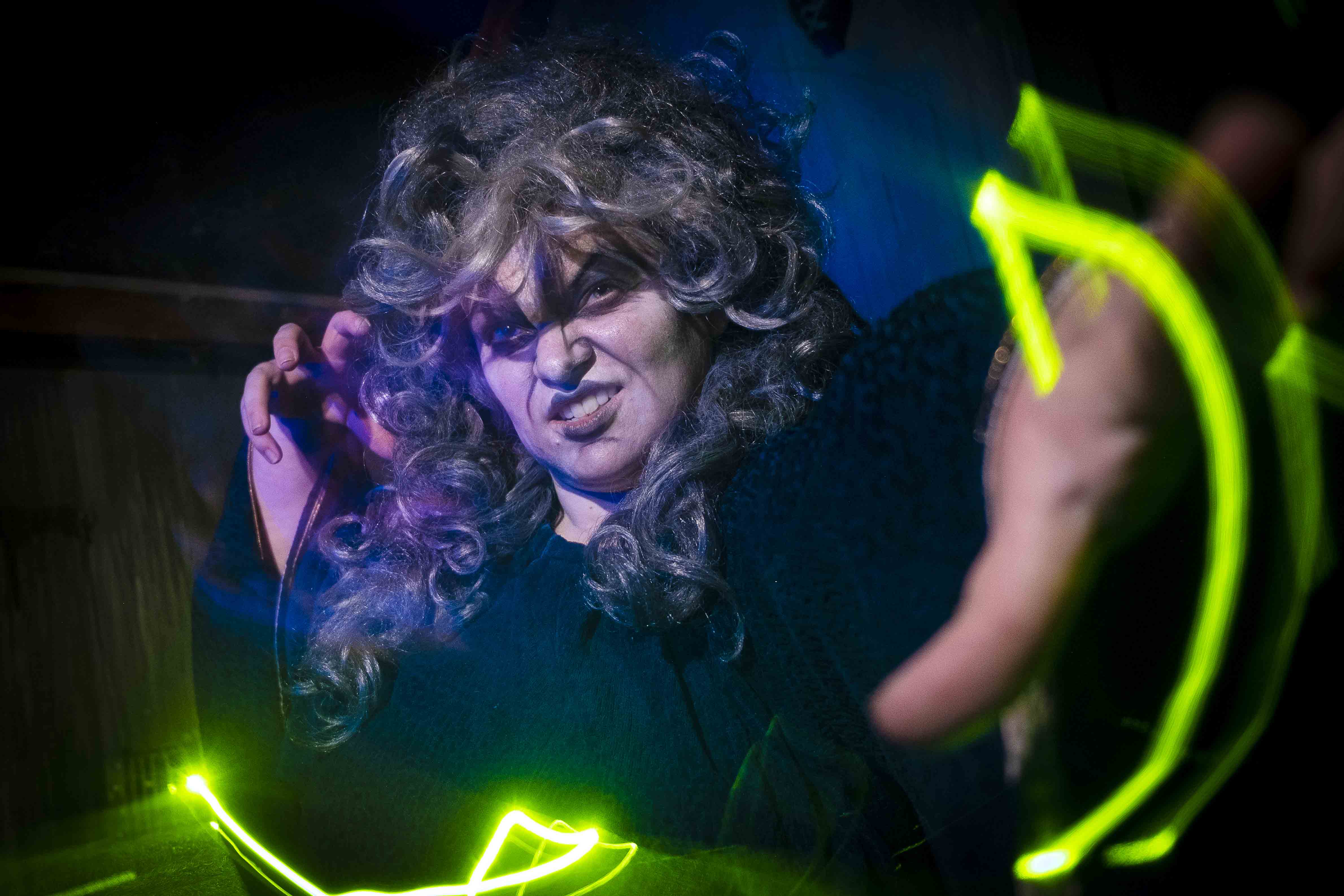 A woman dressed as a witch with long gray hair, scowling, and holding glowing green light trails.
