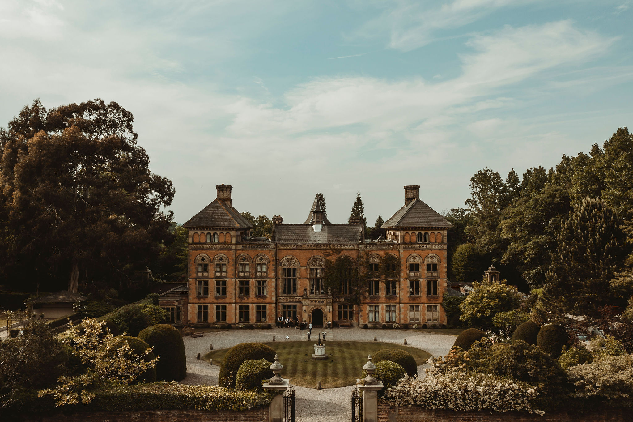 A couple holds hands, standing on a rocky beach under a large cave arch, gazing at the tranquil ocean during sunset. The sky is softly lit in pastel hues.